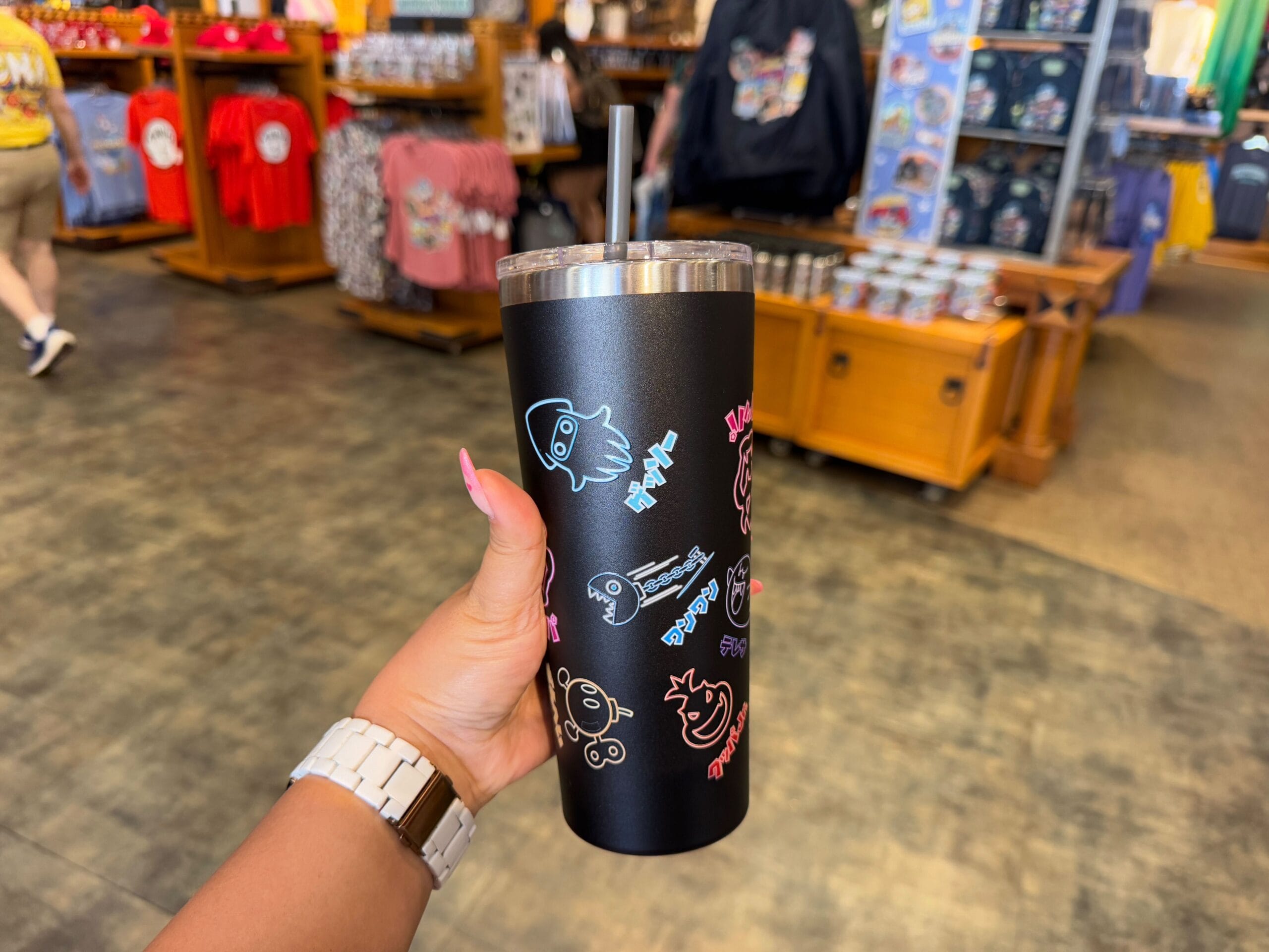 In a store filled with shelves of clothes and intriguing items, a person holds a black tumbler adorned with colorful cartoon animal designs—an exclusive piece from the Epic Universe Merchandise collection at Universal Studios Florida.