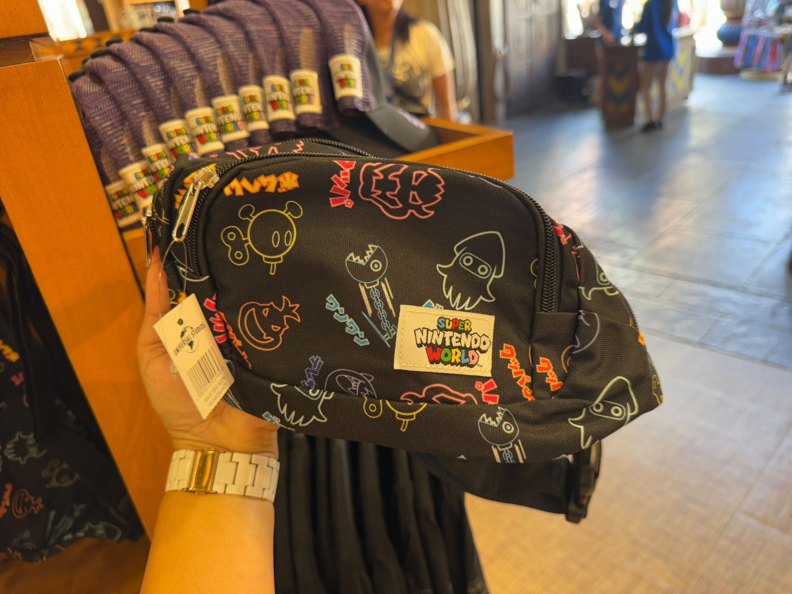 A hand holds a black Nintendo-themed fanny pack, part of the NEW Epic Universe Merchandise collection, decorated with colorful characters and text. More items are displayed in the background, reminiscent of a vibrant day at Universal Studios Florida.