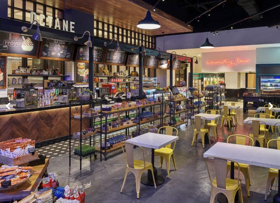 Modern cafe with yellow chairs, white tables, snack shelves, a coffee counter, and a neon sign reading "candy bar" in the back.
