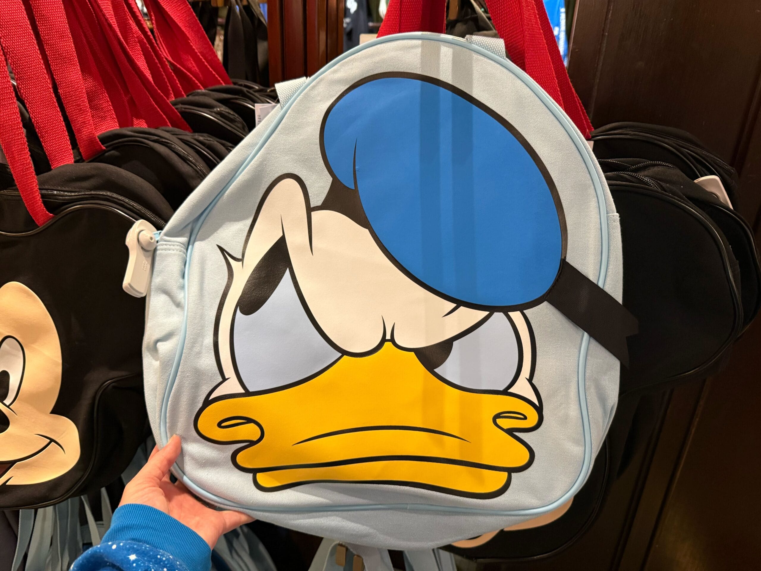 A round tote bag with a grumpy cartoon duck face design, resembling Donald Duck with his blue sailor hat and bright orange beak, stands out against a backdrop of hanging red straps.
