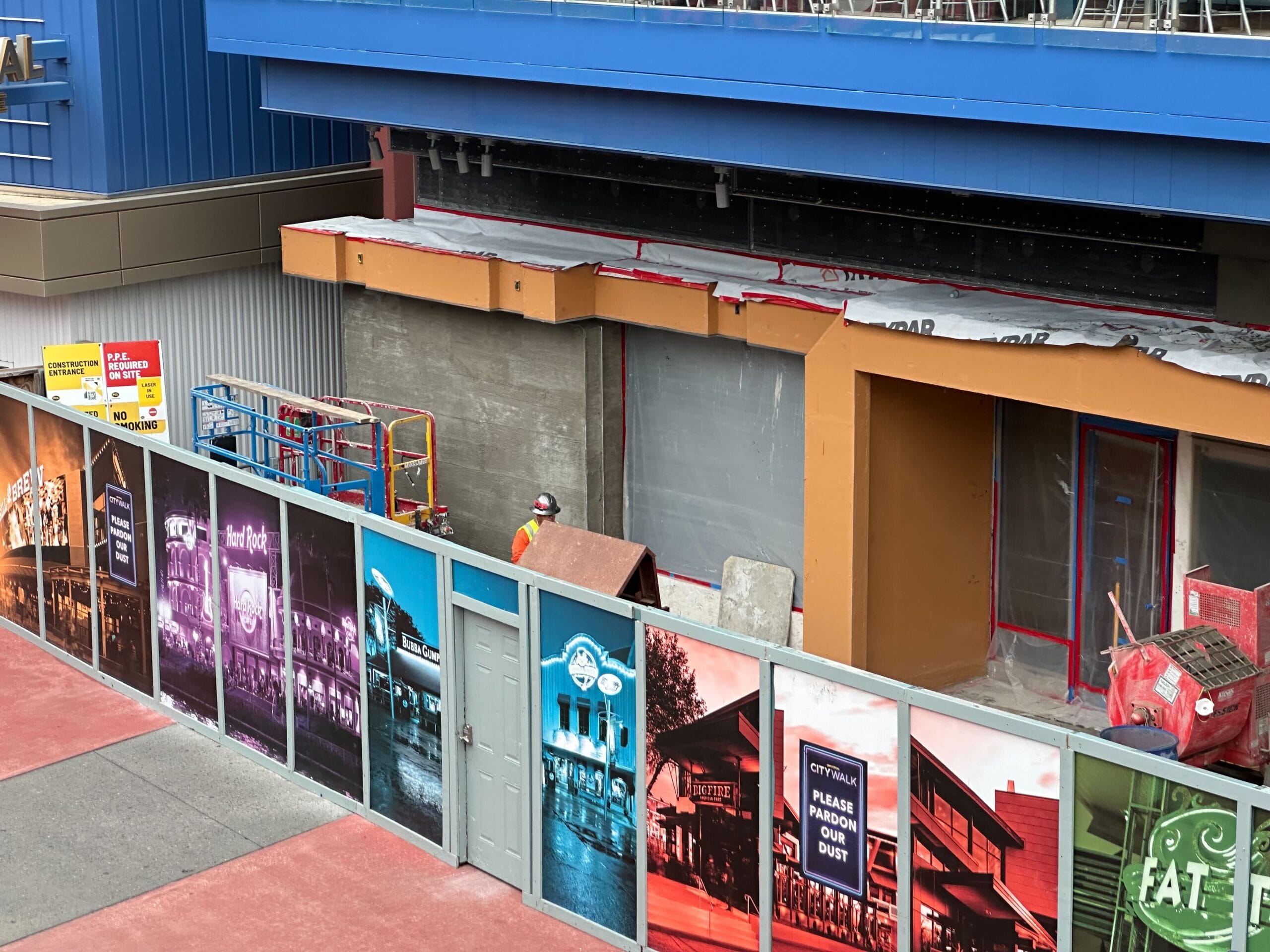 Construction site with a building under renovation, surrounded by colorful panels displaying various images. A worker in a yellow hard hat is present.