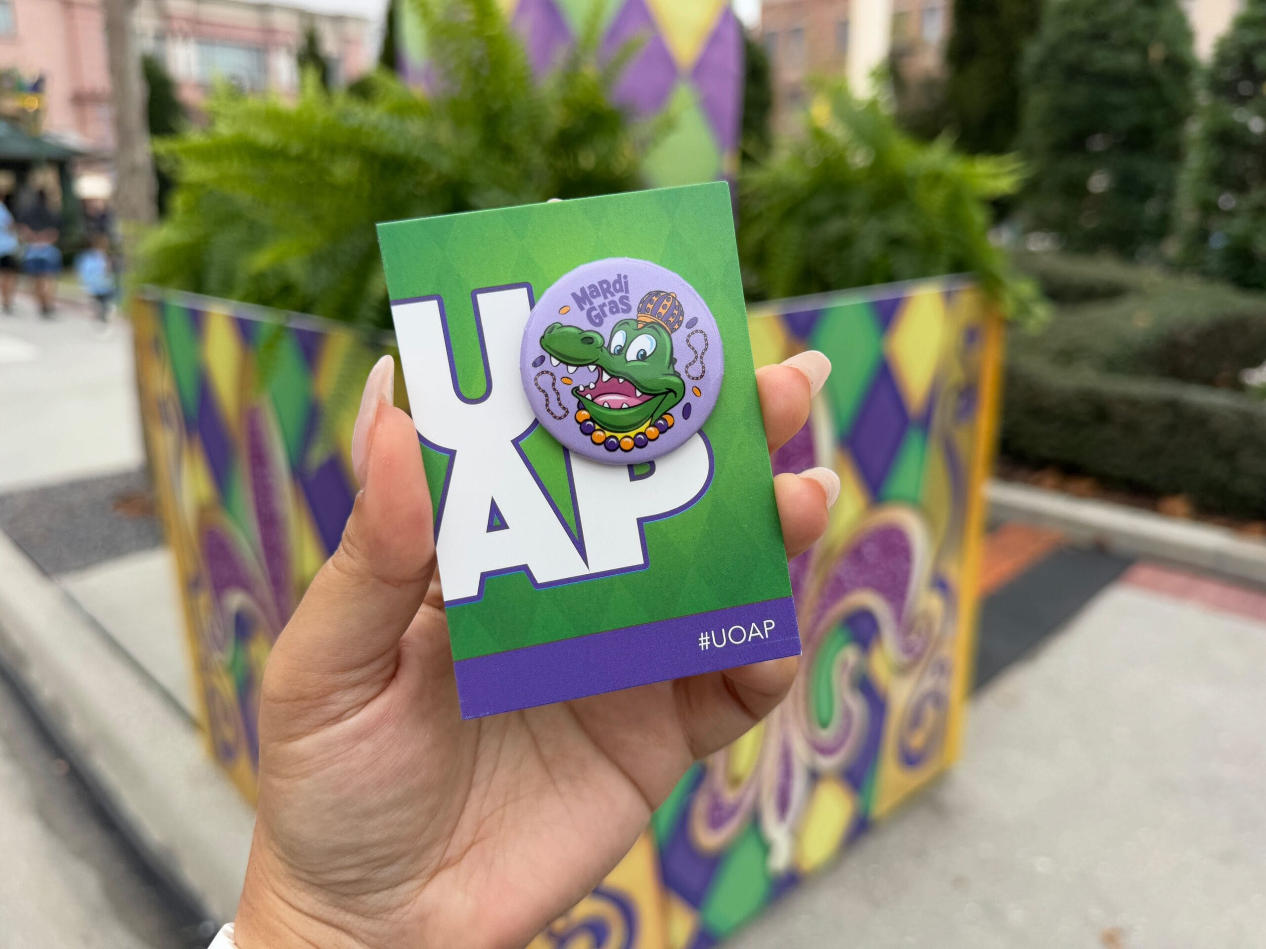 A hand holds a purple and green badge with a cartoon alligator and beads, labeled "Mardi Gras 2025." The backdrop showcases colorful decorations and plants at Universal Orlando Resort, perfectly capturing the vibrant spirit of an Annual Passholder's exclusive celebration.