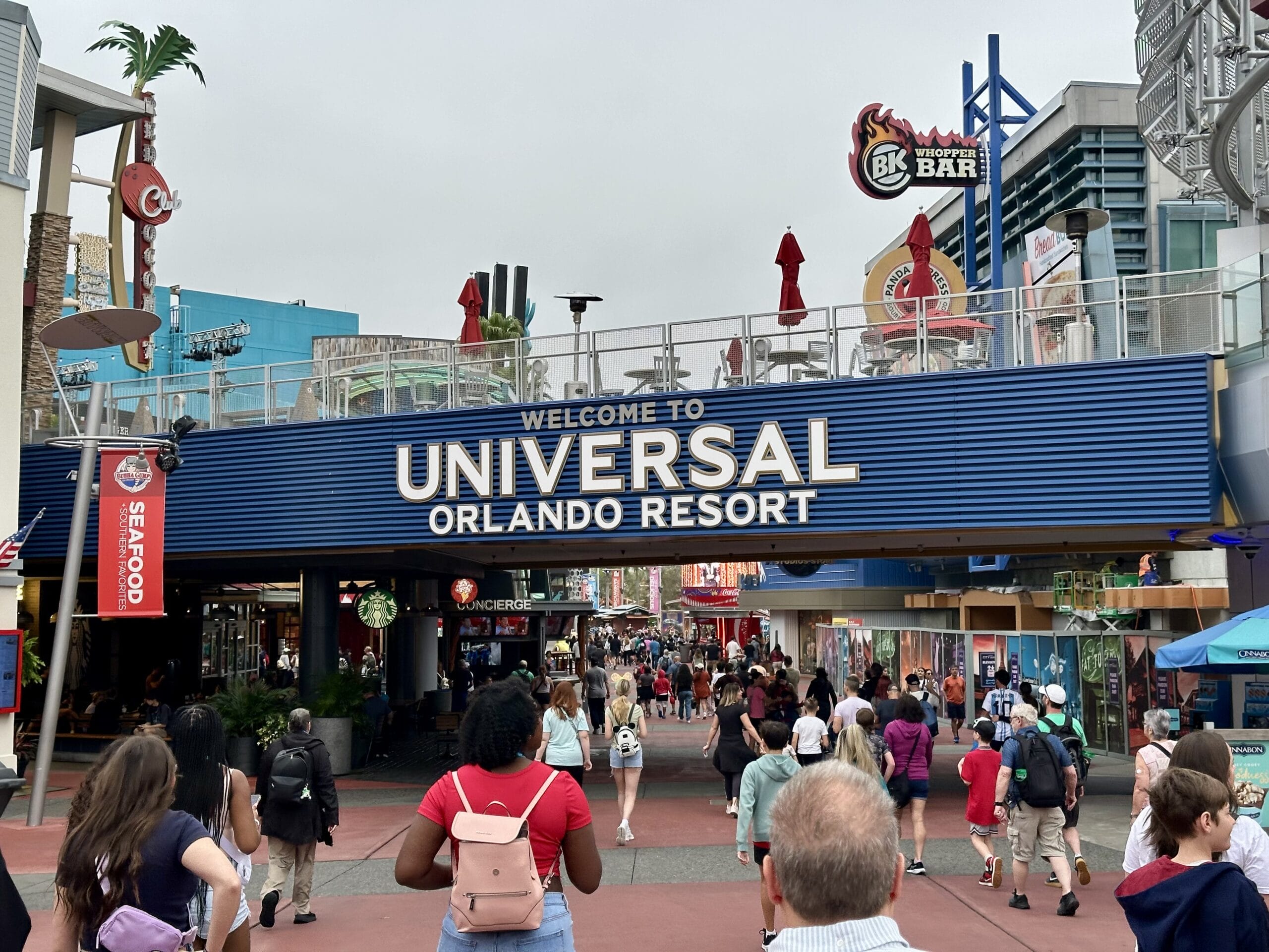 Visitors stroll beneath a welcoming sign that reads "Welcome to Universal Orlando Resort," surrounded by shops and restaurants buzzing with activity. Big sales are everywhere, offering irresistible deals on exclusive EPIC Universe merchandise for a limited time.