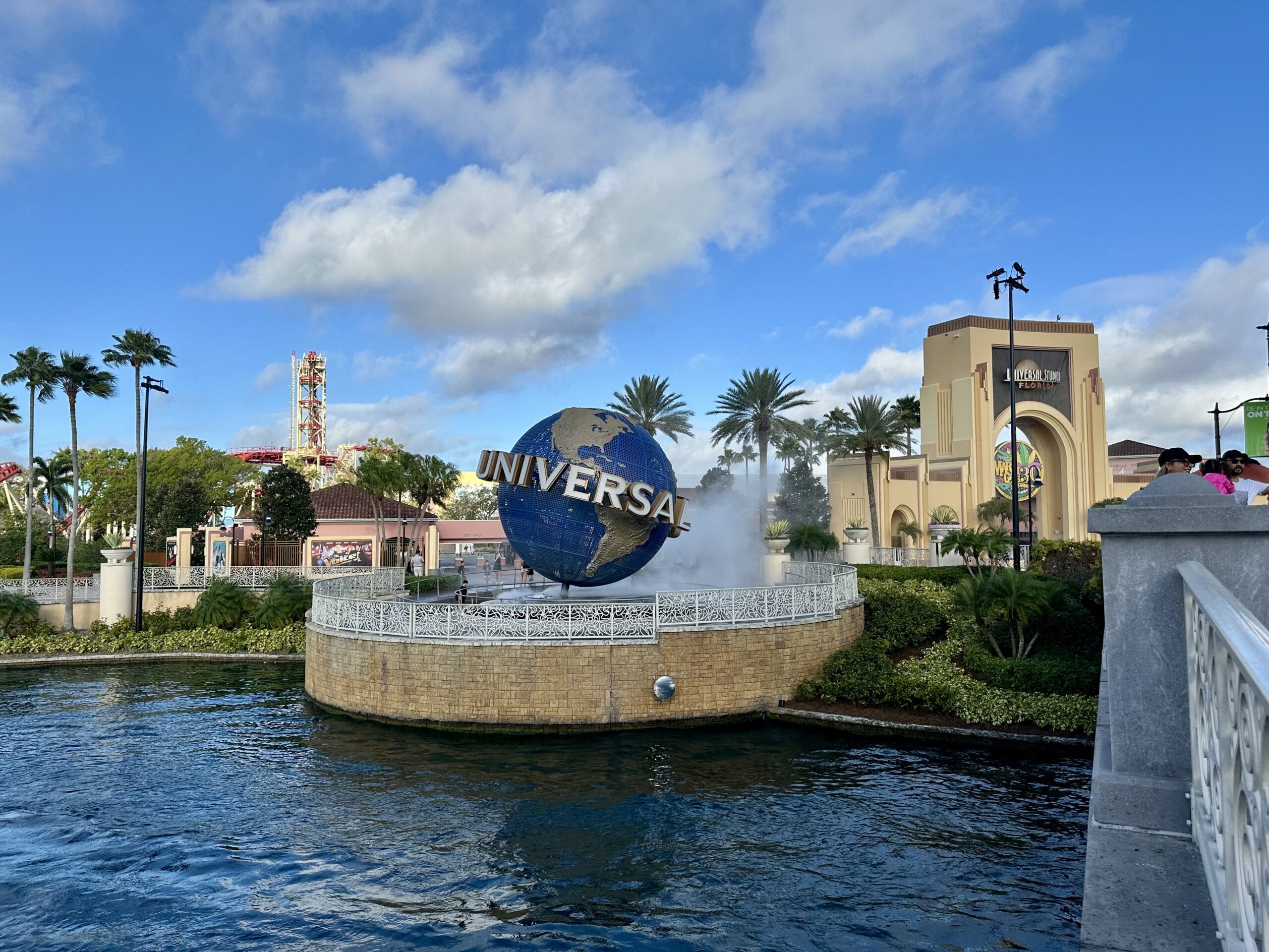 A globe with "Universal" stands on a misty fountain, surrounded by palm trees and park buildings beneath a partly cloudy sky. It's the perfect backdrop for your Universal Orlando photo report during Mardi Gras festivities.