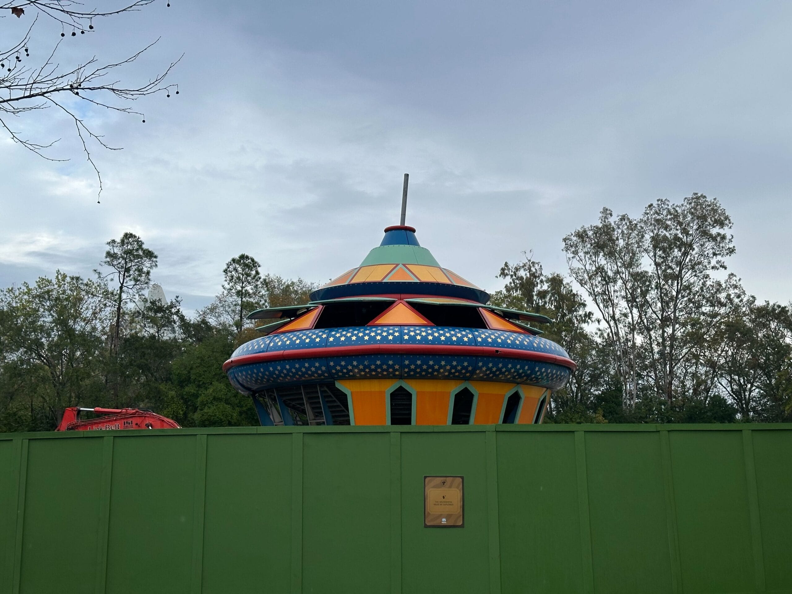 A colorful UFO-shaped structure is partially visible behind a green construction fence, with trees and a cloudy sky in the background.