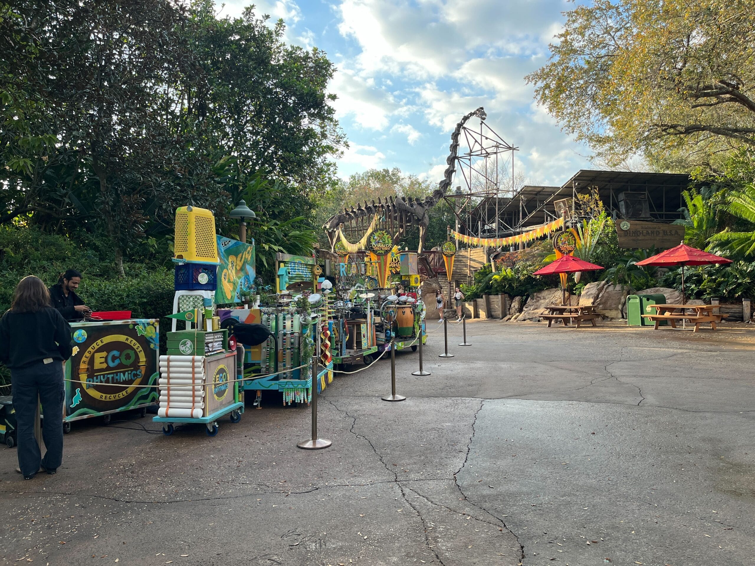 The vibrant performance location, known as "Eco Cart," features colorful vending carts surrounded by trees, red umbrellas, and picnic tables. Under a cloudy sky, a large skeleton display adds intrigue to this lively scene in the heart of our very own Animal Kingdom.