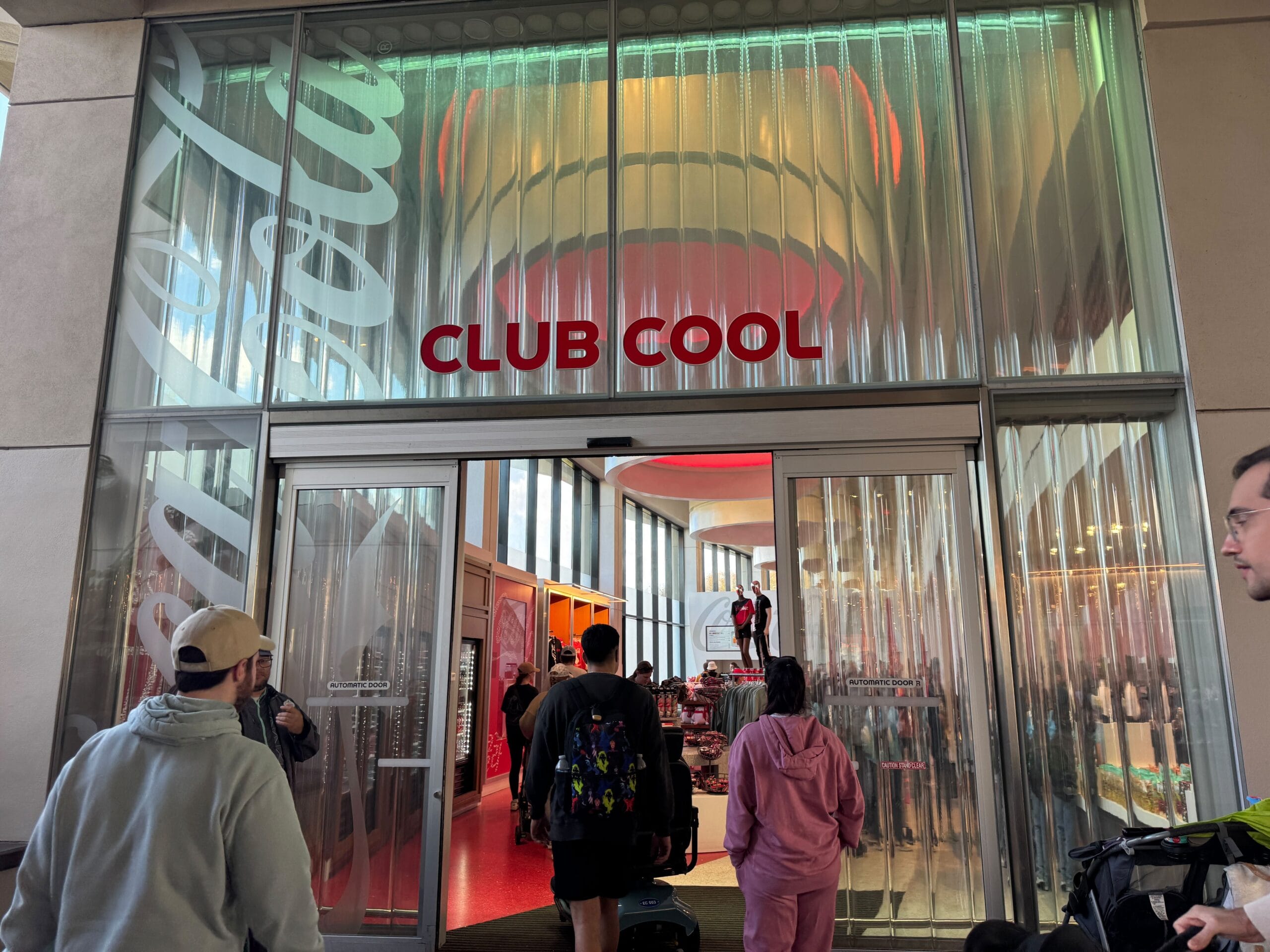 People entering Club Cool, a Coca-Cola themed store with a glass facade. Red and white signage visible.