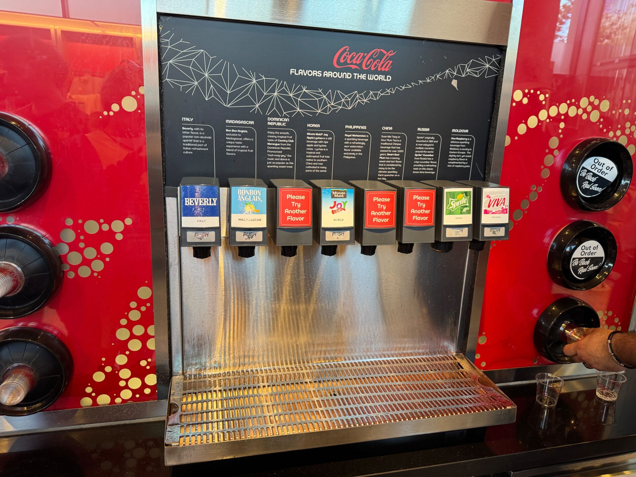 Coca-Cola global flavors soda dispenser with various drink options, including Beverly, Sparberry, and others. A person is using the machine, and there are several empty cups on the drip tray.