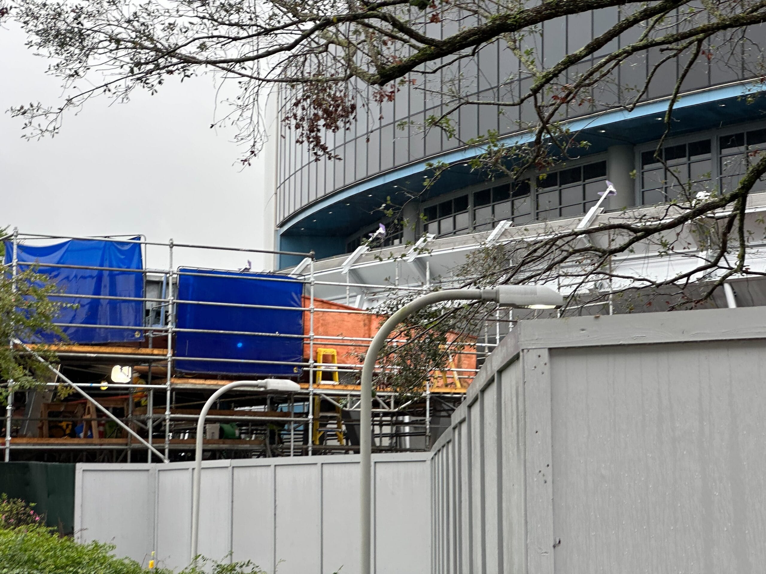 A building under construction, enshrouded in blue and orange tarps with scaffolding hinting at progress, stands tall. In the foreground, a tree and streetlights defy an overcast sky, reminiscent of an EPCOT vision.