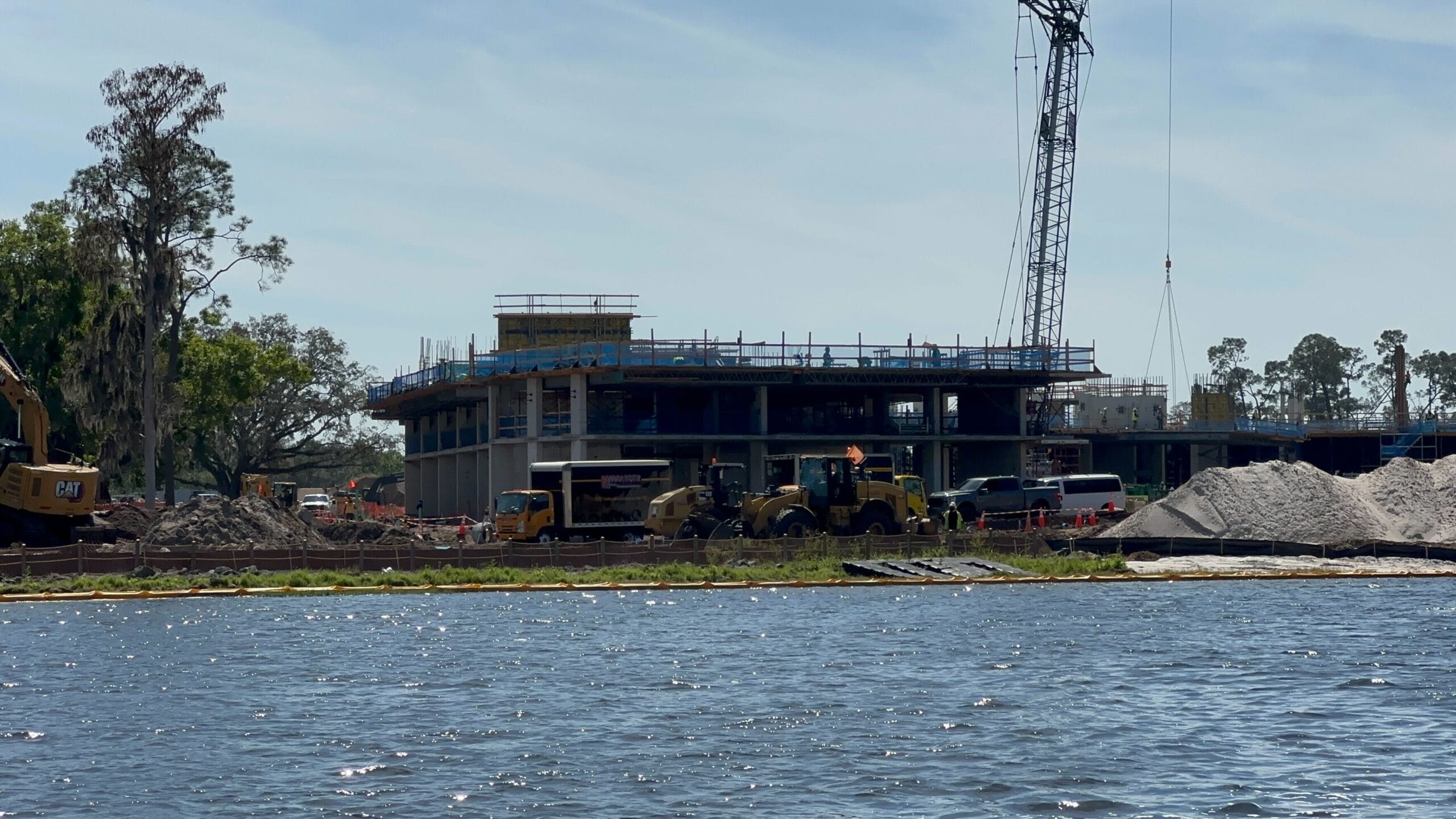 Beside the tranquil lakeshore, a bustling construction site emerges with cranes and machinery, shaping a partially built structure under a clear sky.