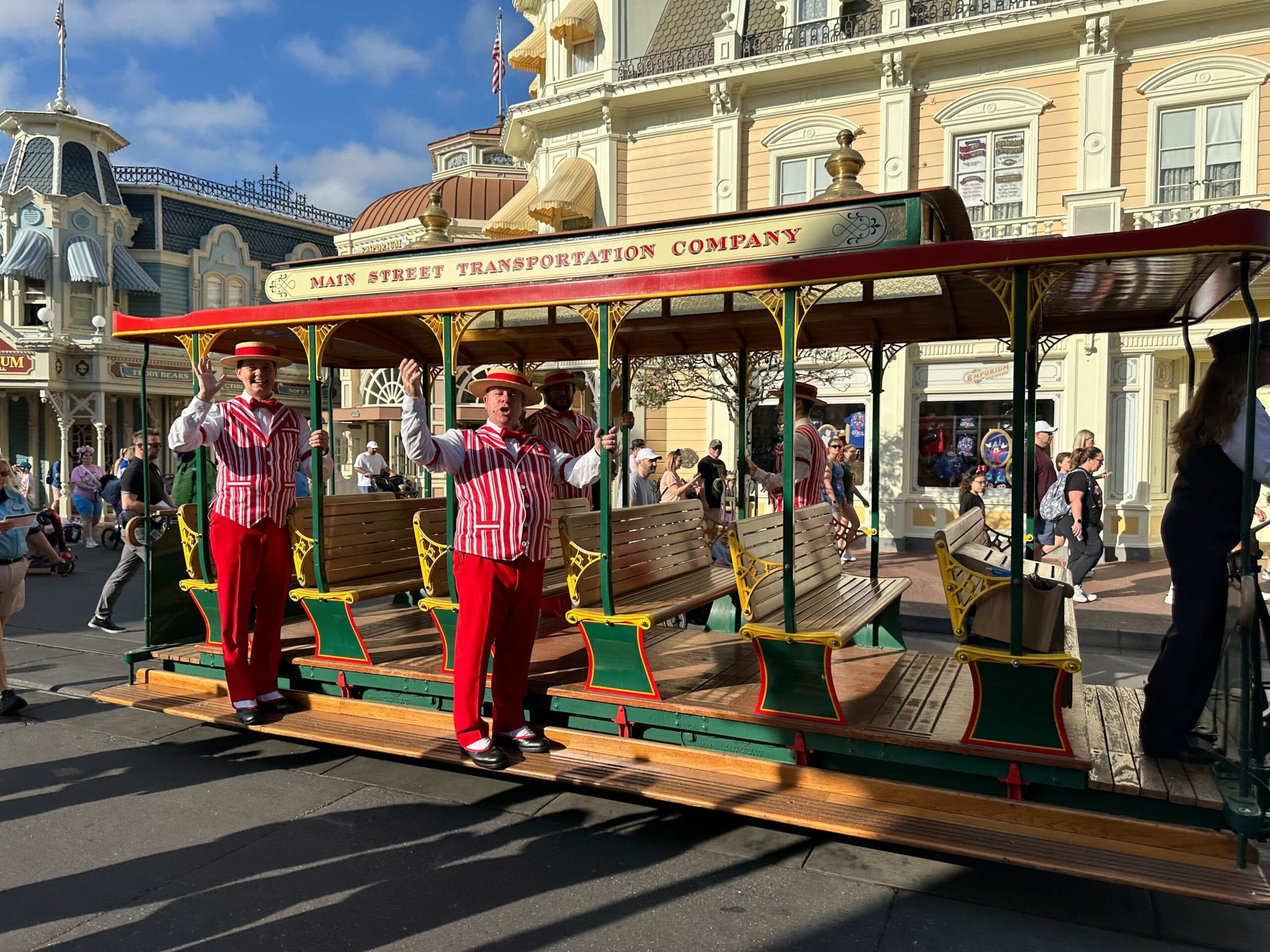 A vintage-style trolley with "Main Street Transportation Company" on the sign rolls down a street in Magic Kingdom. Two uniformed staff members in red and white wave cheerfully, reminiscent of the Dapper Dans, as people stroll by in the background.
