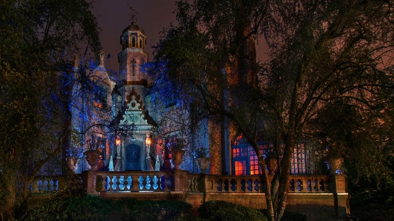 A dimly lit, ornate building with a blue and red glow, surrounded by trees, casts a mysterious allure reminiscent of the Haunted Mansion at Walt Disney World when viewed at night.