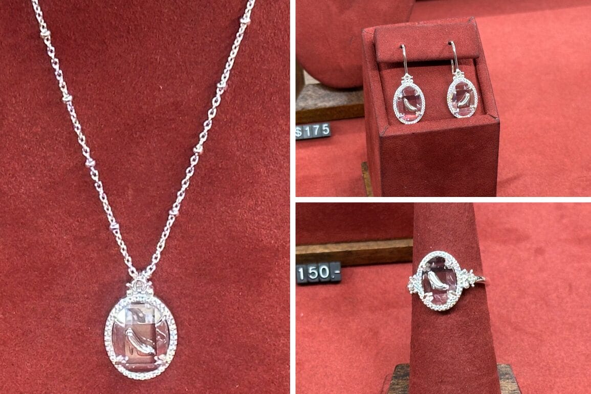 This Rebecca Hook silver jewelry set, displayed on luxurious red velvet, features an exquisite oval pendant necklace, matching earrings, and a ring. Adorned with clear stones and intricate detailing, this collection celebrates Cinderella's 75th Anniversary. Price tags are visible.