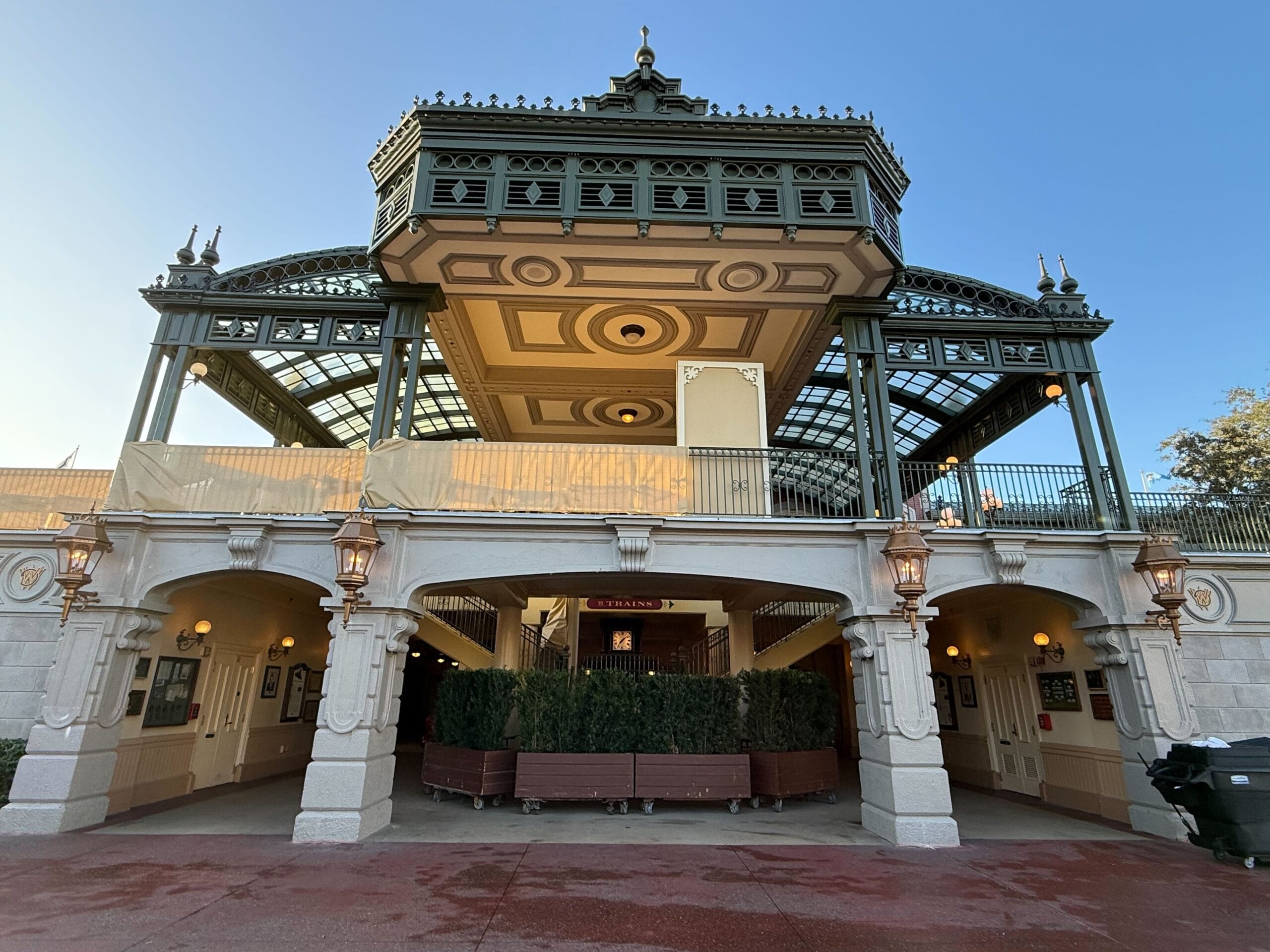 Train station entrance with decorative overhang, glass panels, and ornate lamps. The structure features a mix of beige and green accents, with a clear blue sky in the background.