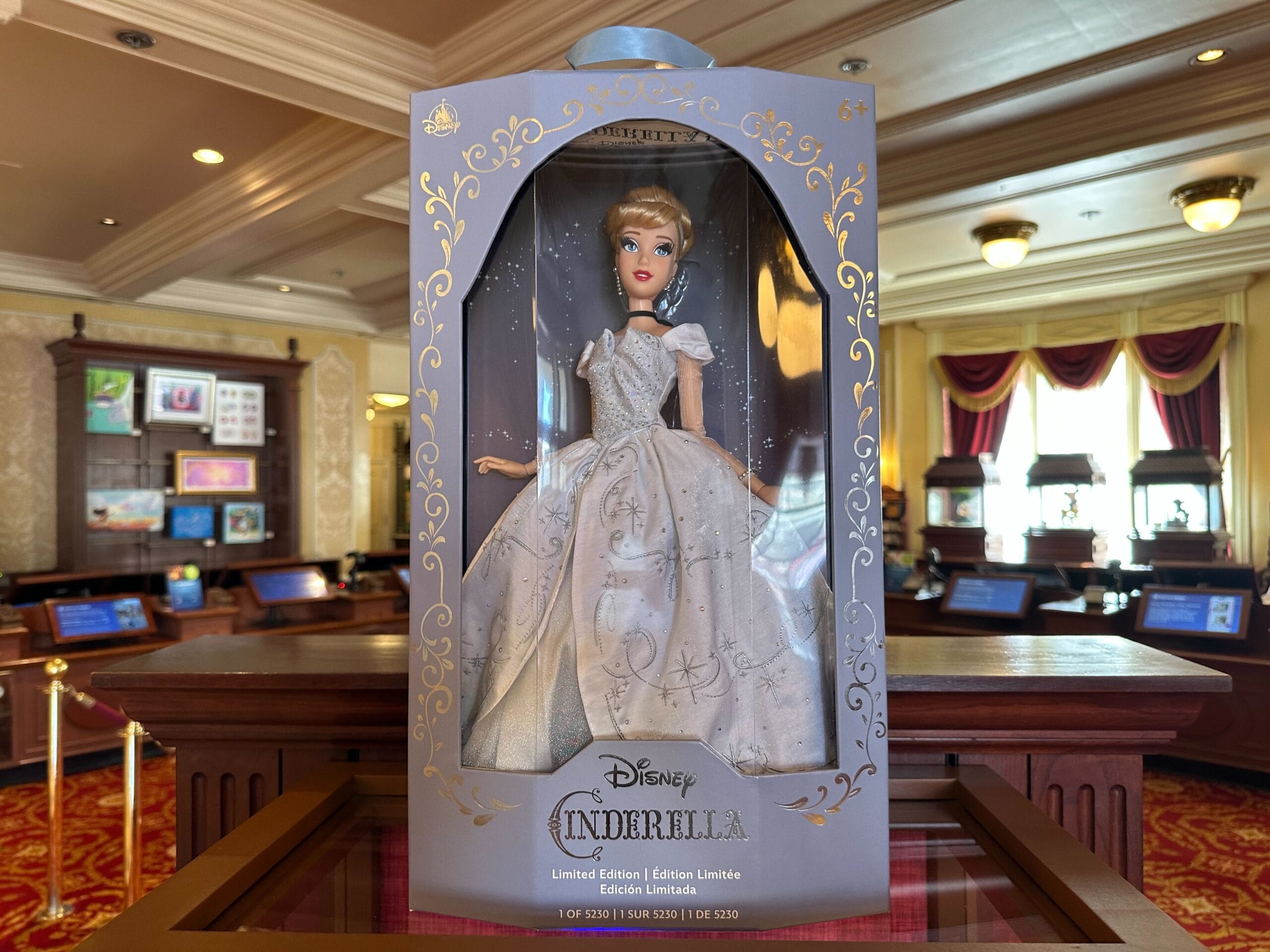 A Limited-Edition Cinderella doll in an elegant dress is displayed in a decorative box. The transparent front reveals the doll, celebrating the Cinderella 75th Anniversary. It's showcased in a well-lit store with wooden shelves, reminiscent of Walt Disney World's magical allure.