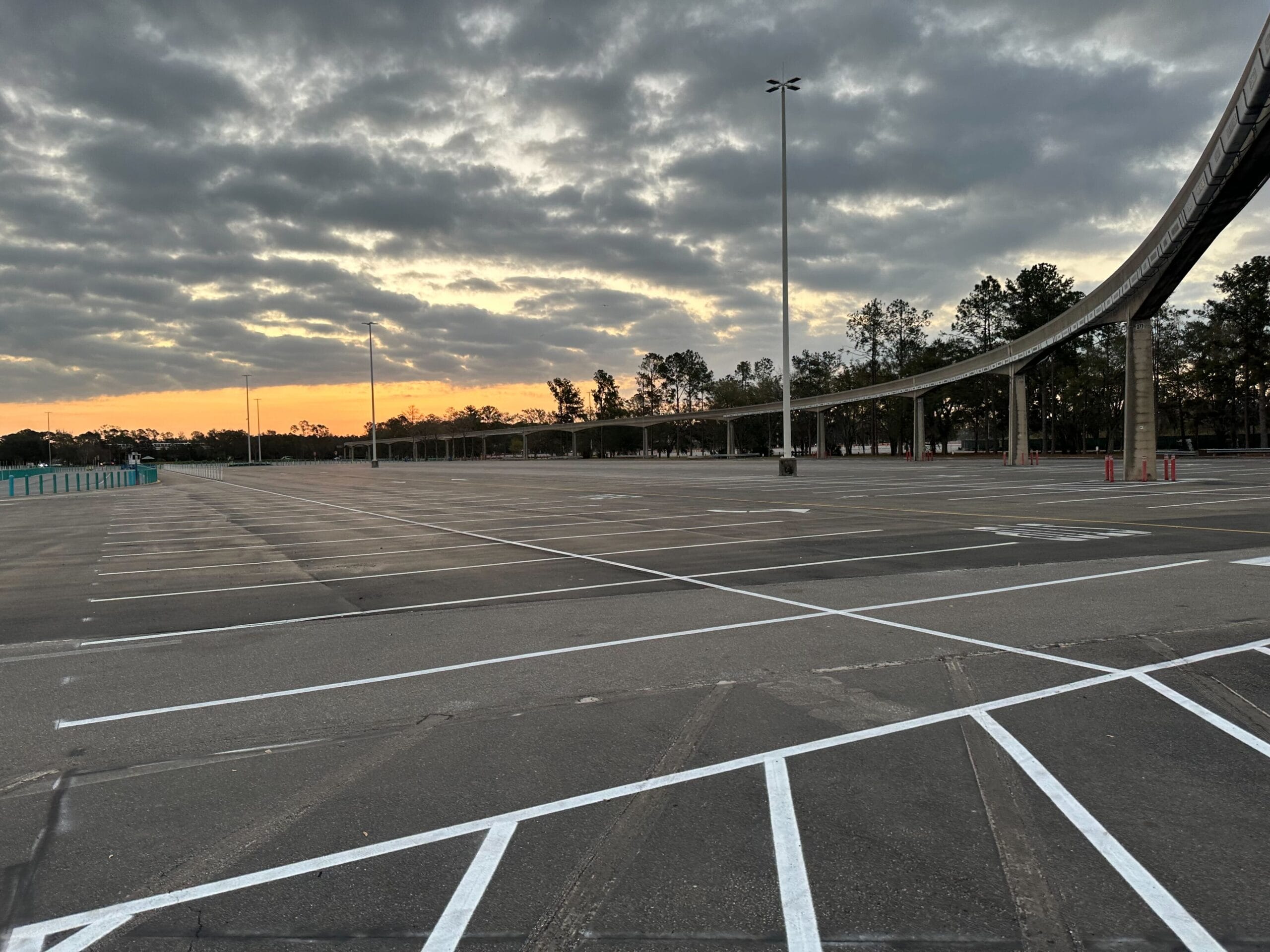 An empty parking lot at sunset with overhead lights and monorail track visible. The sky is partly cloudy.