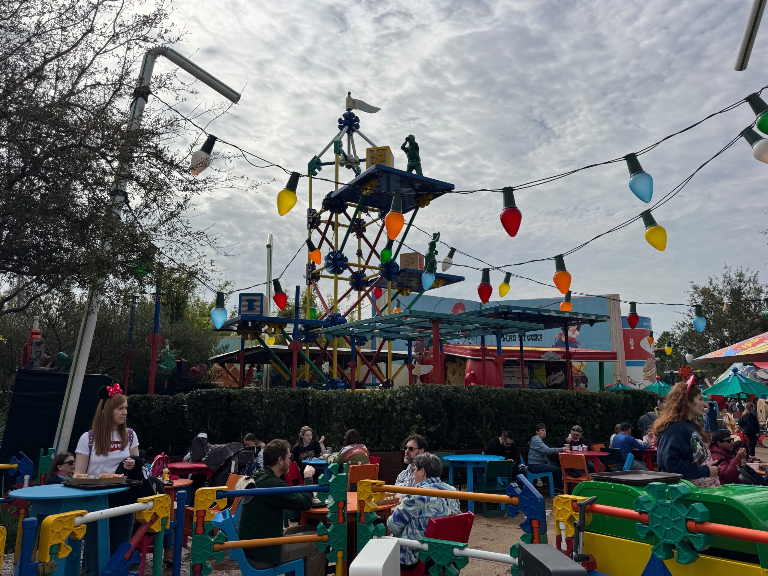 People sitting at outdoor tables in a theme park are enjoying the lively ambiance. Colorful decorations and string lights hang overhead, near the iconic Woody's Lunch Box. A large structure resembling a toy set adds to the whimsical atmosphere.