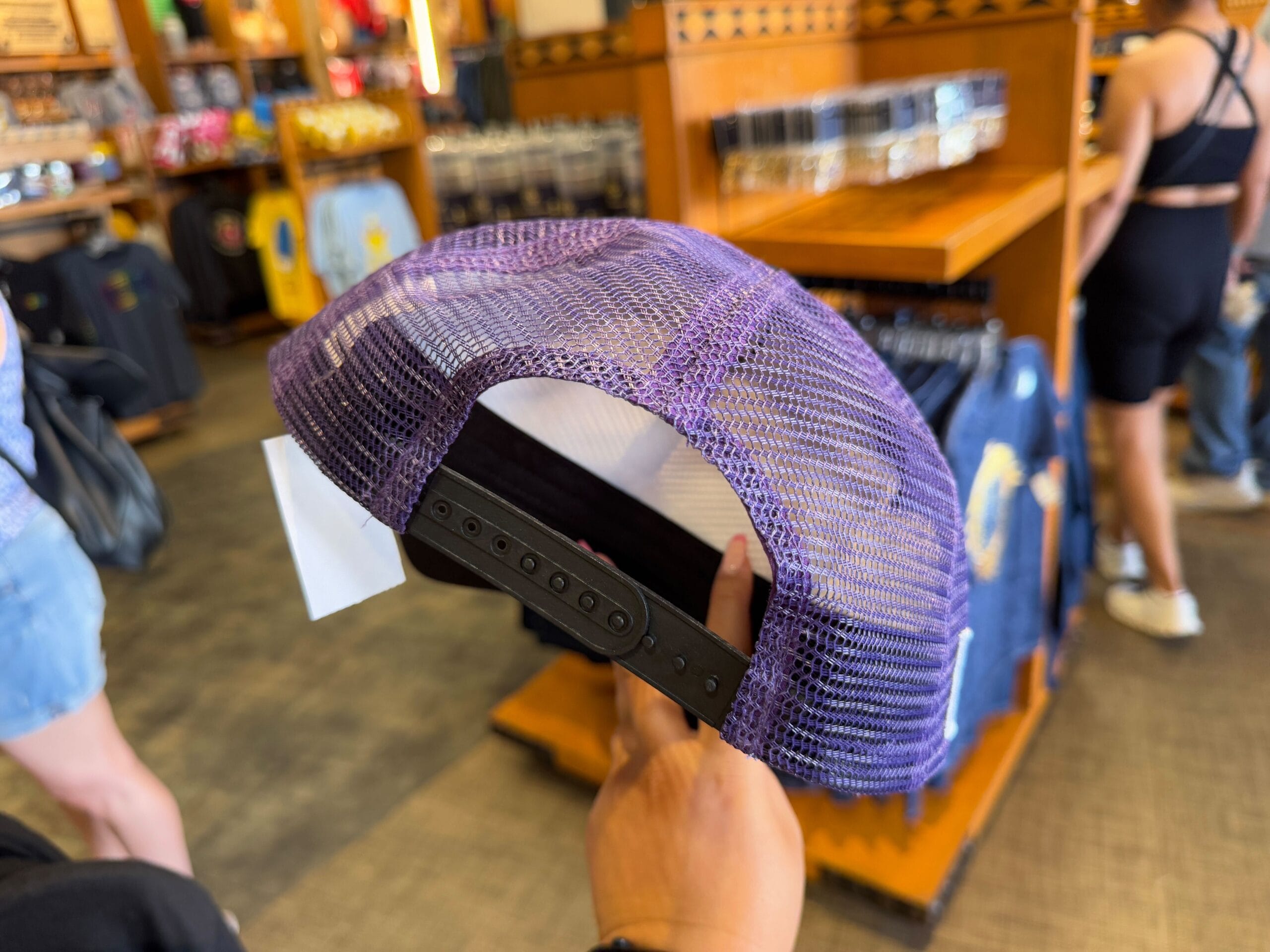 A person holds an Epic Universe Merchandise purple mesh trucker hat inside a store at Universal Studios Florida, with wooden shelves and various apparel in the background.
