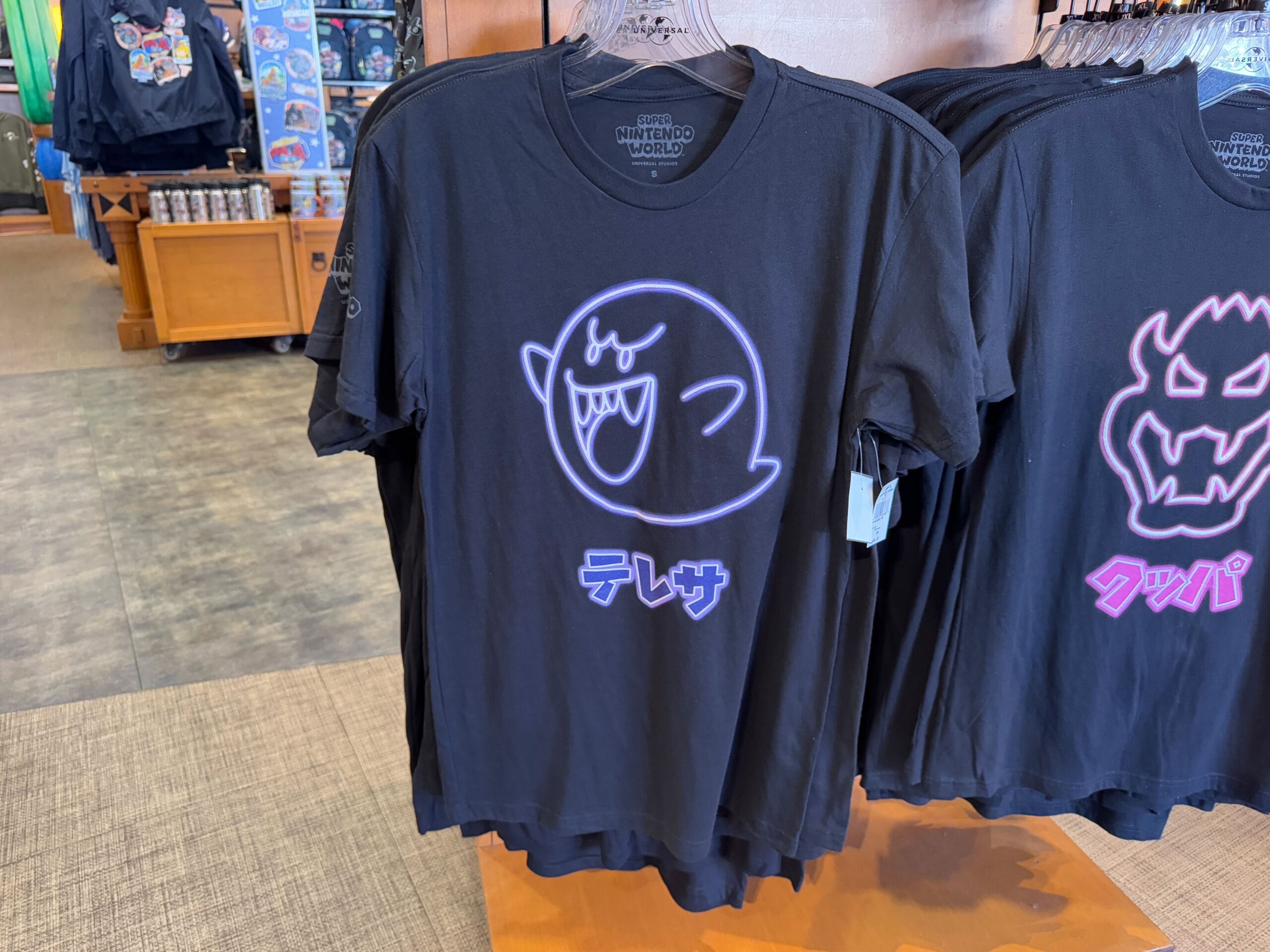Black T-shirt featuring a glowing ghost graphic and Japanese text, displayed on a rack in the Epic Universe Merchandise section at Universal Studios Florida.