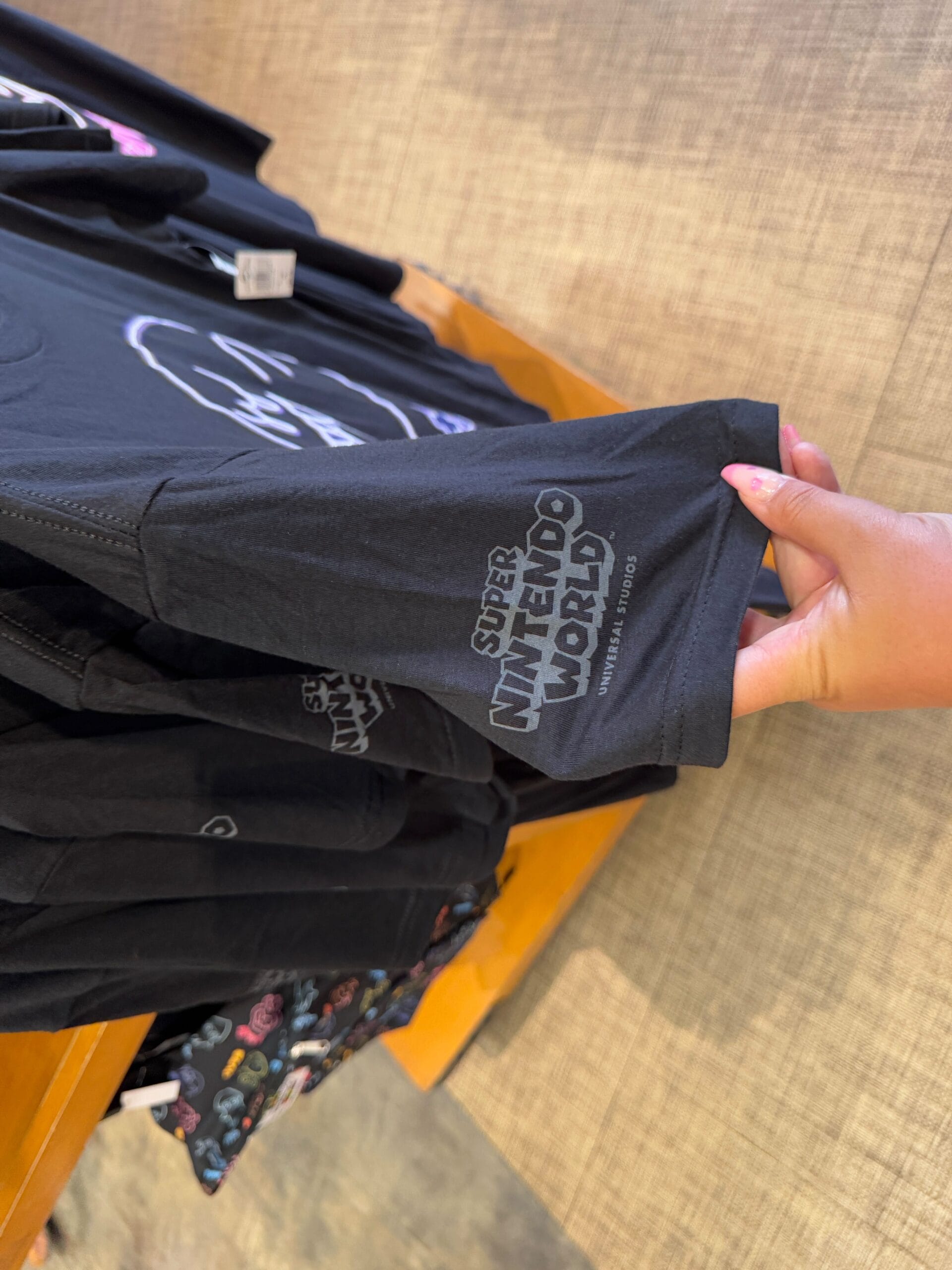 A person holds a black "Super Nintendo World" T-shirt sleeve, displayed on a wooden rack with more black T-shirts—a stylish piece of Epic Universe Merchandise found at Universal Studios Florida.
