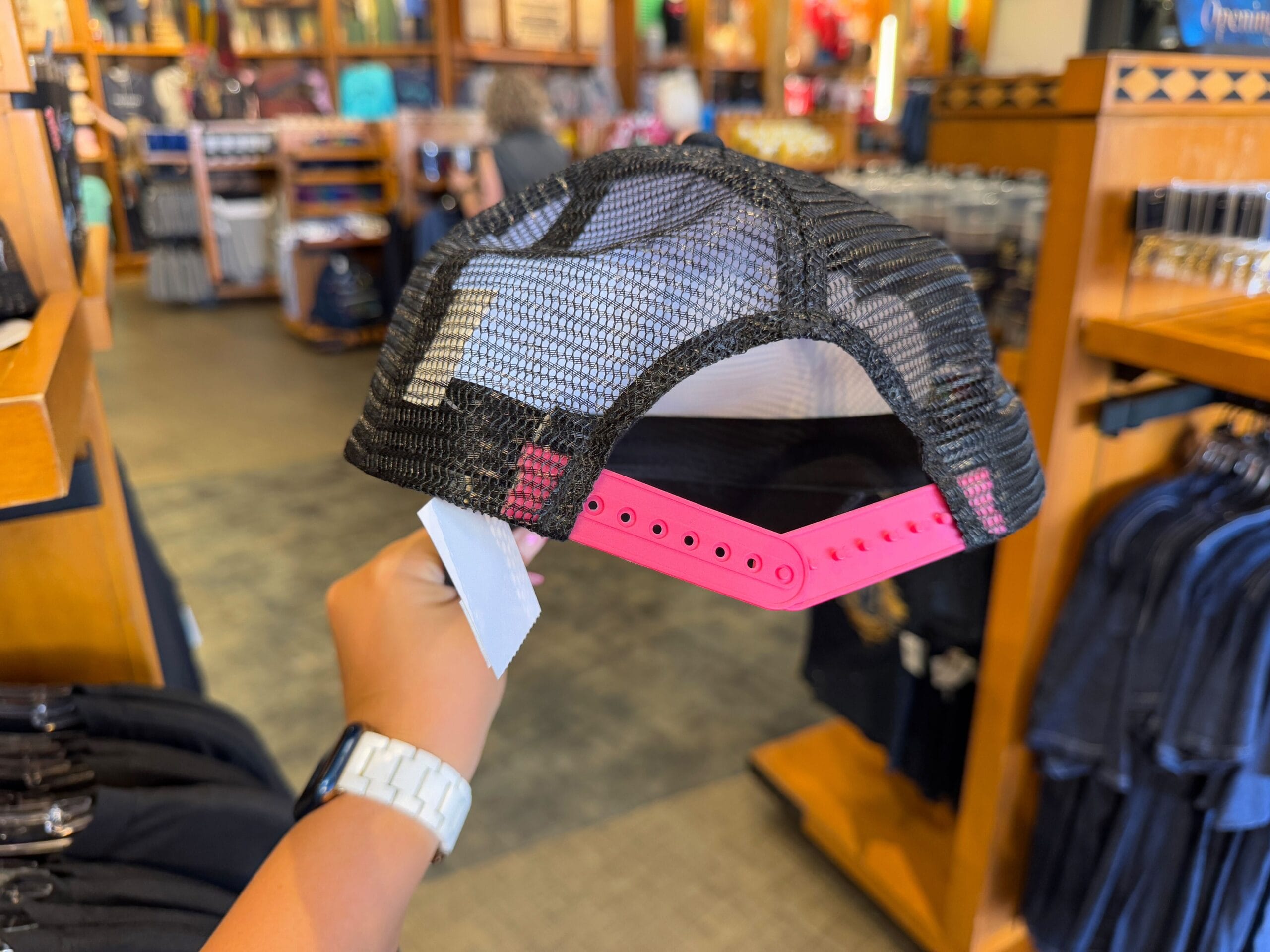 Inside a store, a person holds a black mesh cap featuring Epic Universe Merchandise from Universal Studios Florida, complete with a bright pink adjustable strap.