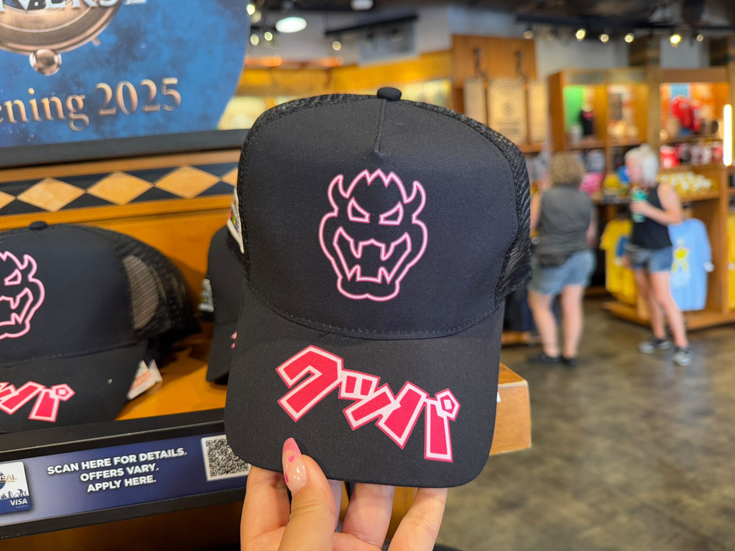 Black cap with a pink Bowser logo and Japanese text, available as part of the Epic Universe Merchandise at Universal Studios Florida. Displayed in a store with other themed items in the background.