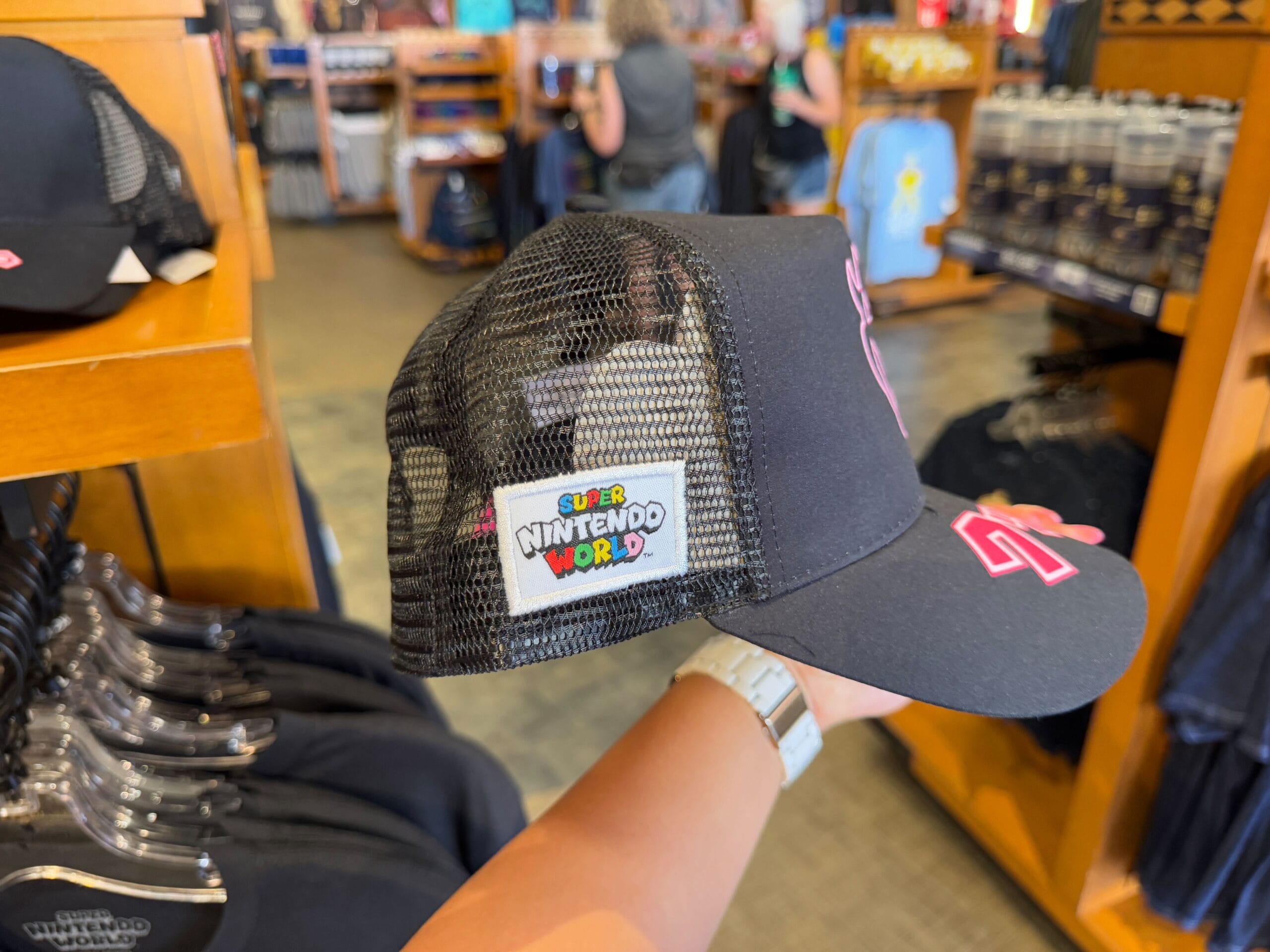 In a bustling store setting at Universal Studios Florida, a hand displays a black mesh-back cap adorned with a pink design on the front and an exclusive "Super Nintendo World" patch on the side—an exciting addition to your Epic Universe merchandise collection.