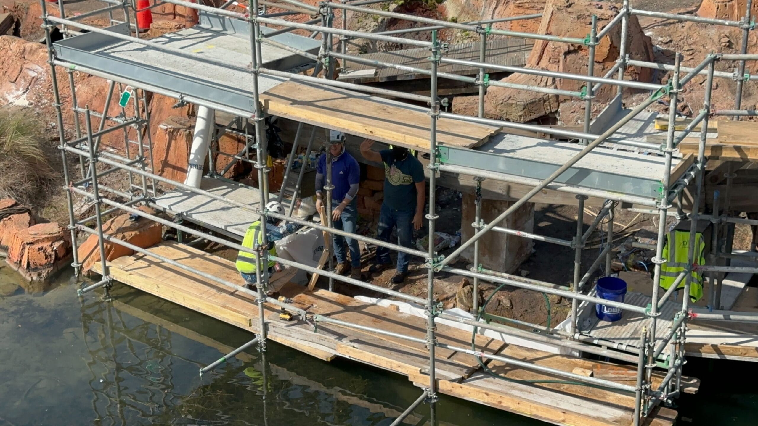 Crew members stand on scaffolding above the water, diligently engaged in a construction task. The scaffolding is supported by the rugged, rocky backdrop reminiscent of Big Thunder Mountain Railroad.
