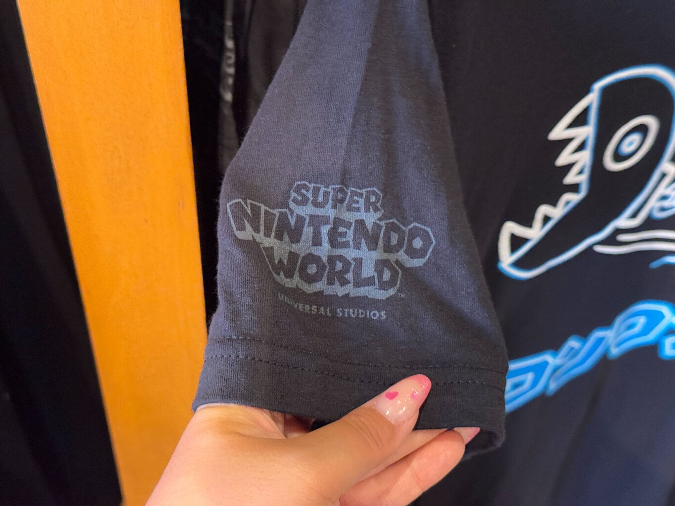 A hand holds a black t-shirt sleeve showcasing a "Super Nintendo World" logo with "Universal Studios" text beneath, a perfect piece of Epic Universe Merchandise from Universal Studios Florida.