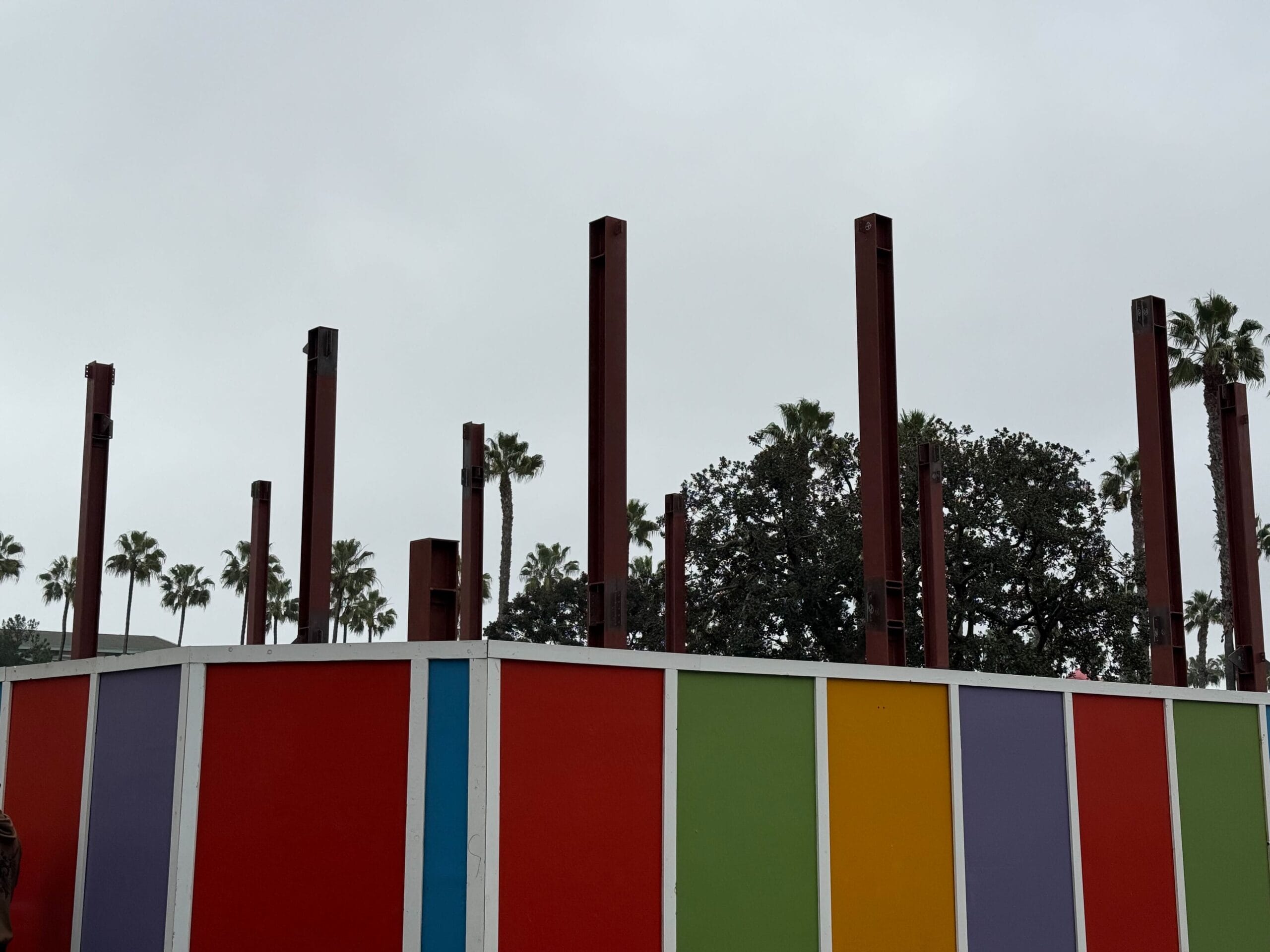 A colorful fence with vertical metal beams protruding above it, set against a cloudy sky and palm trees in the background, hints at construction near Downtown Disney's vibrant atmosphere.