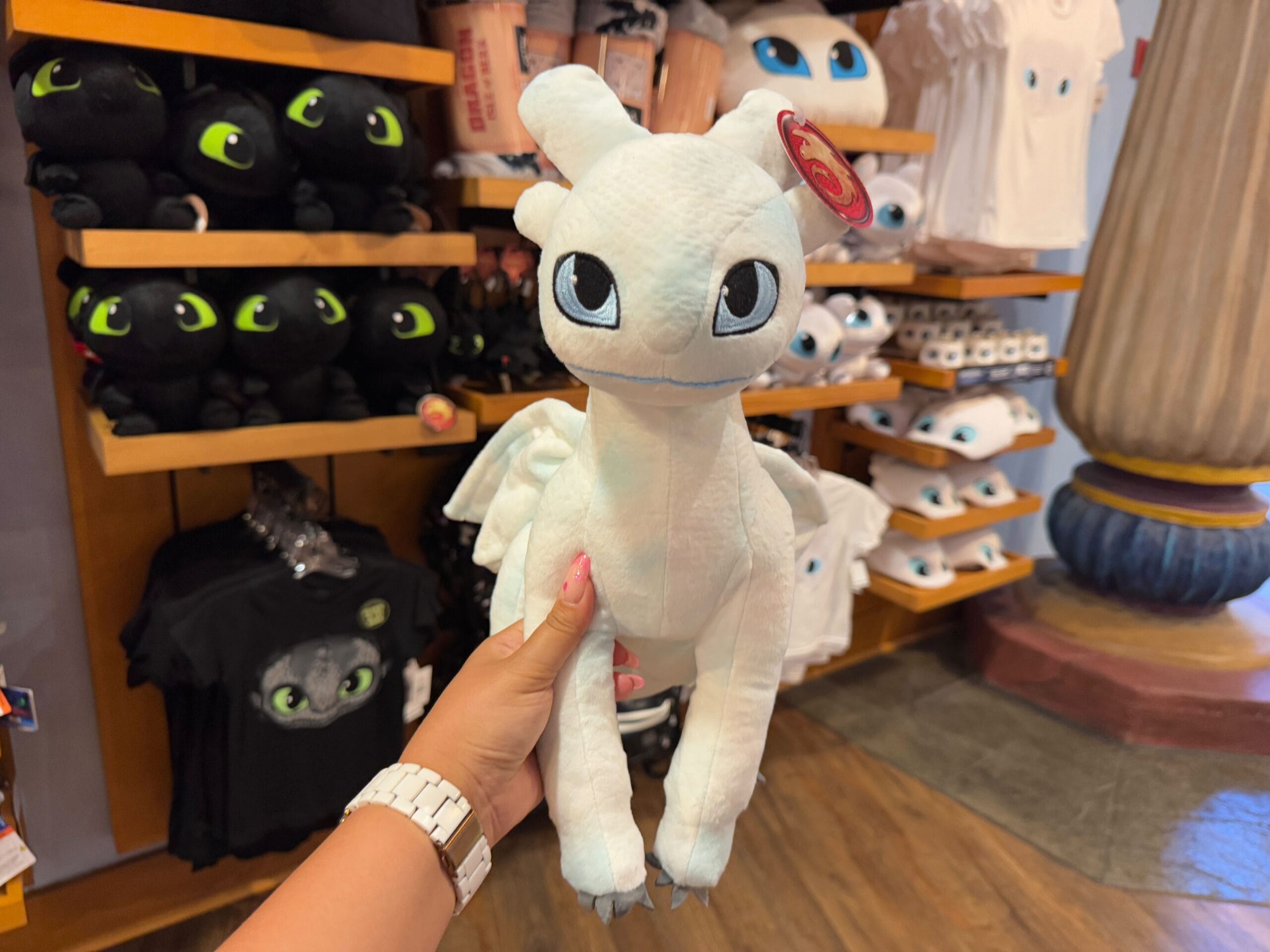 In a store filled with dragon-themed wonders, a person holds a white dragon plush toy, surrounded by shelves brimming with similar treasures and Epic Universe Merchandise from Universal Studios Florida.