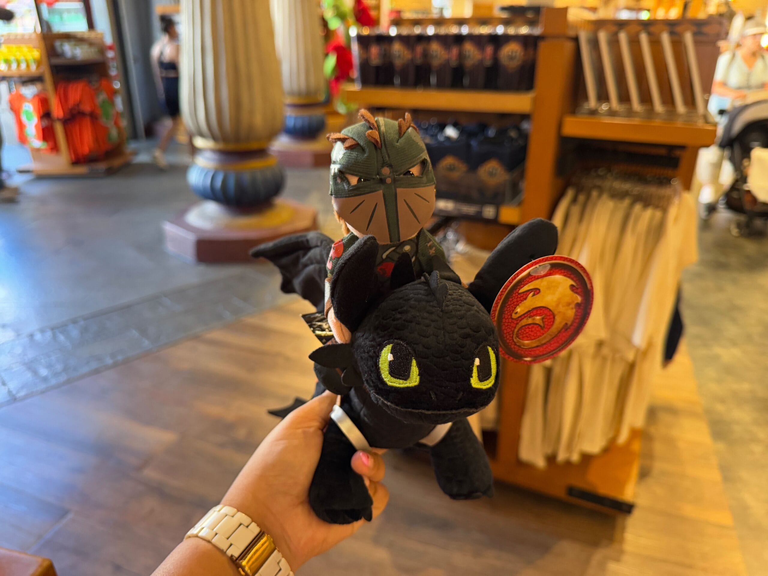 A hand holds a plush toy of a black dragon with large eyes, featuring a small armored figure riding on its back. Clothing and Epic Universe Merchandise from Universal Studios Florida are displayed in the background.