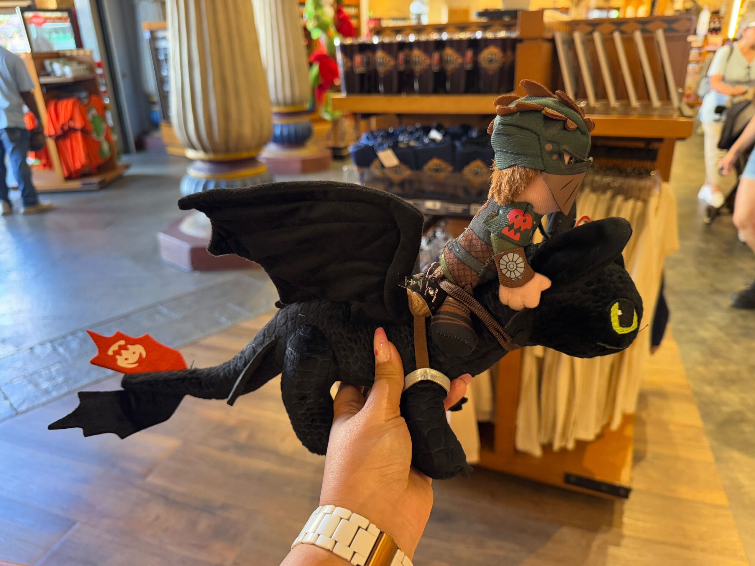 A plush toy of a rider in armor sits atop a black dragon with a red tail emblem, held by someone wearing a watch in the Epic Universe Merchandise store at Universal Studios Florida.