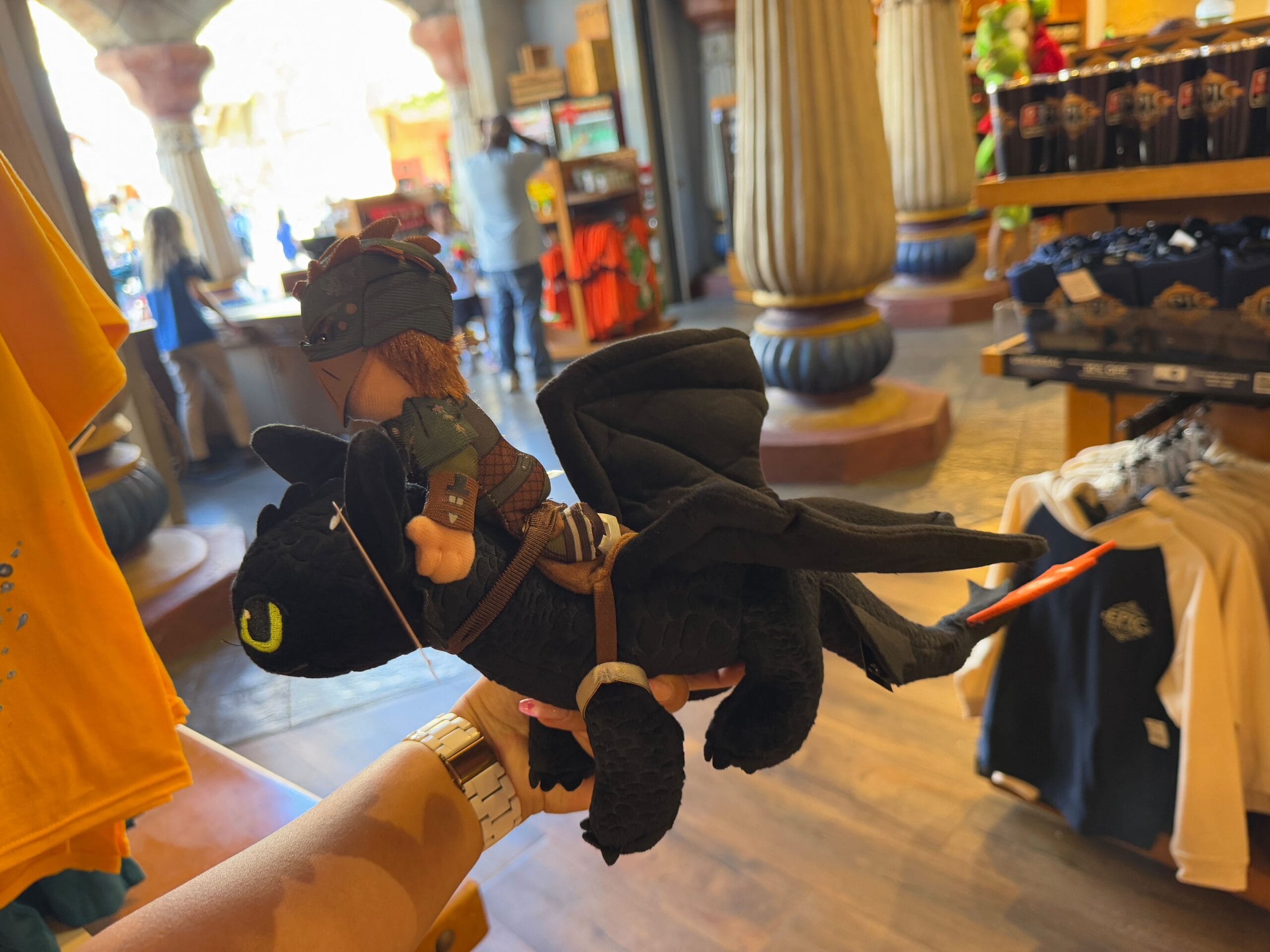 In a store bustling with Epic Universe Merchandise, a hand displays a toy of a small figure riding a black dragon, clothing and shelves forming the backdrop.