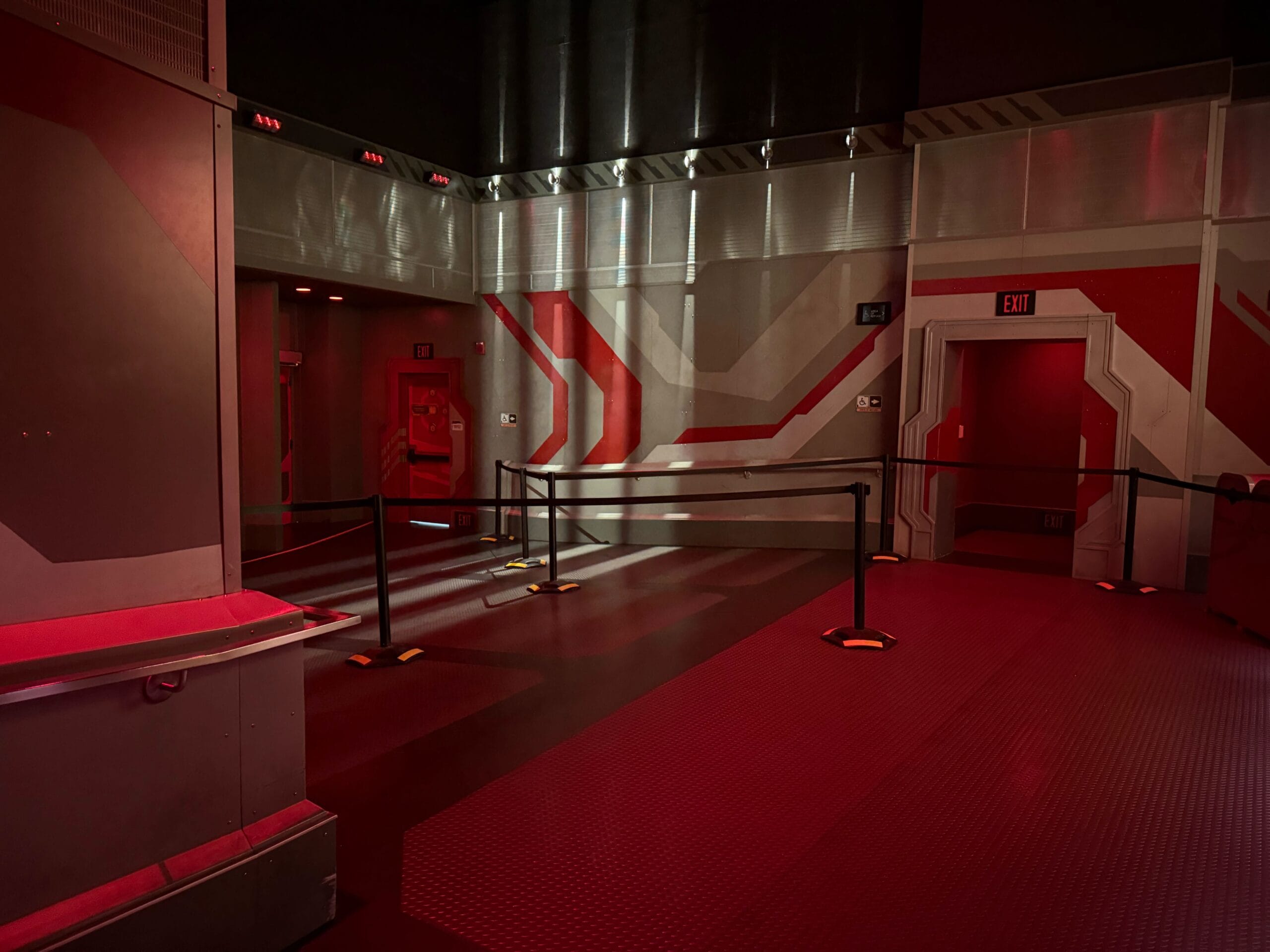 A red-lit room with a cosmic, futuristic design features metallic walls and flooring. A doorway and an exit sign are visible, as black barriers form a line pathway, almost as if anticipating a standby debut for Guardians of the Galaxy: Cosmic Rewind.