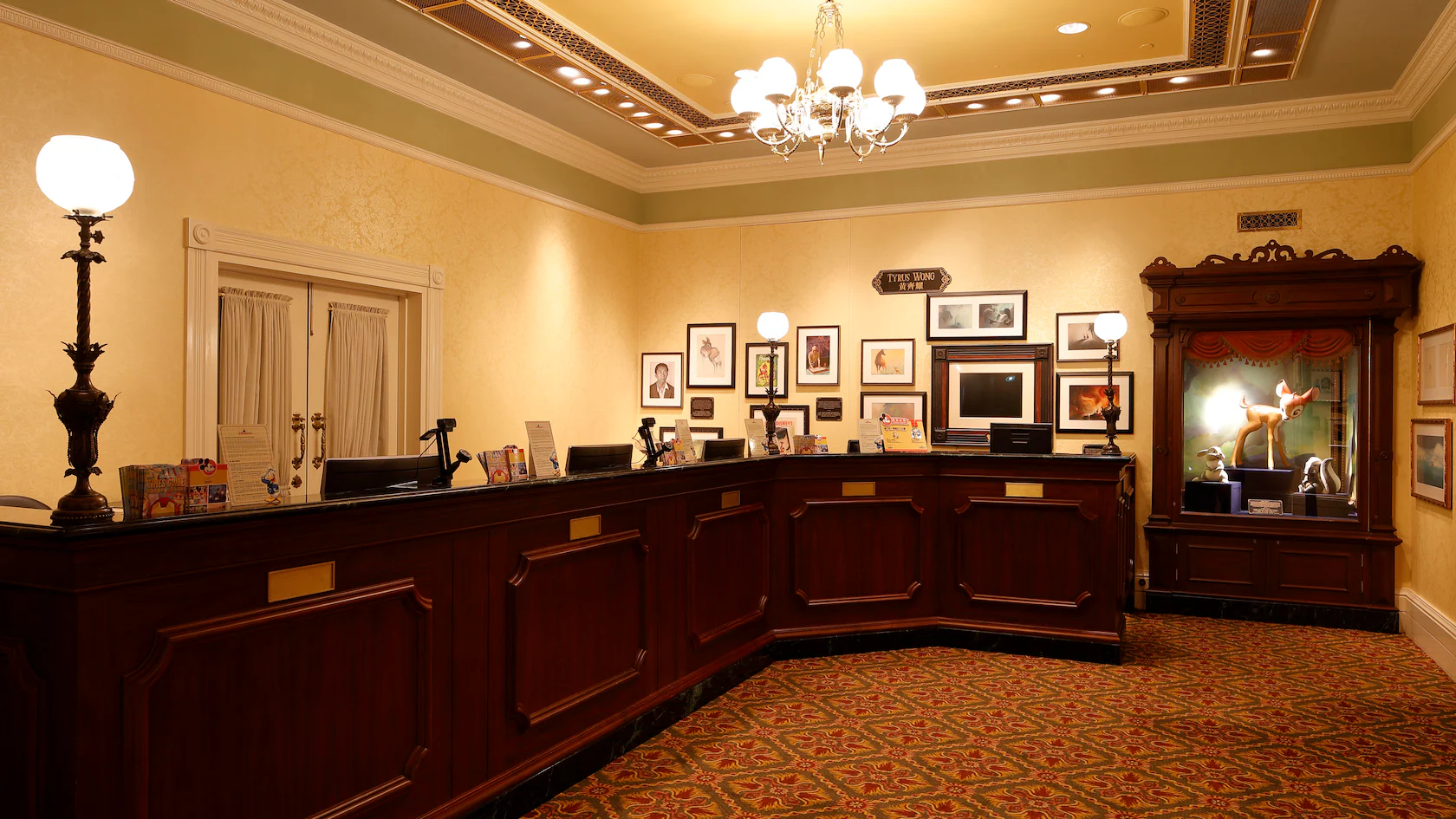 The hotel reception area boasts a wooden desk, an ornate ceiling, and framed pictures that add a touch of art. A statue display graces the corner, bringing to life the art of animation in its enchanting form.