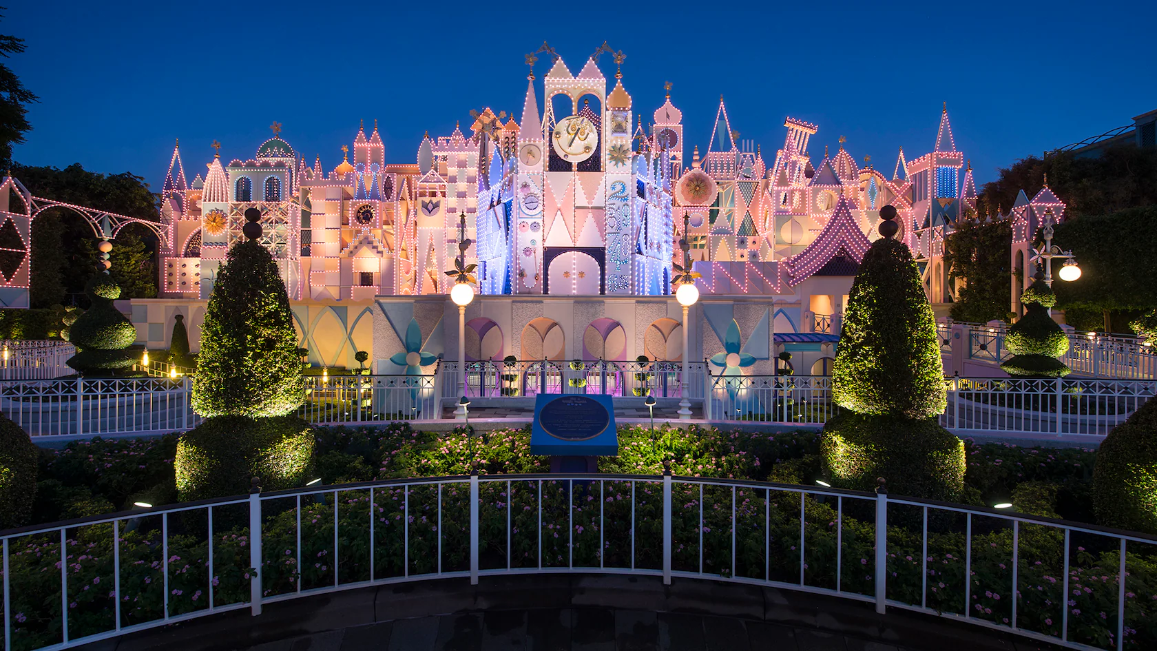 The "it's a small world" attraction features an illuminated, ornate facade and clock tower, surrounded by manicured shrubs and flowers, all set against an enchanting evening sky.