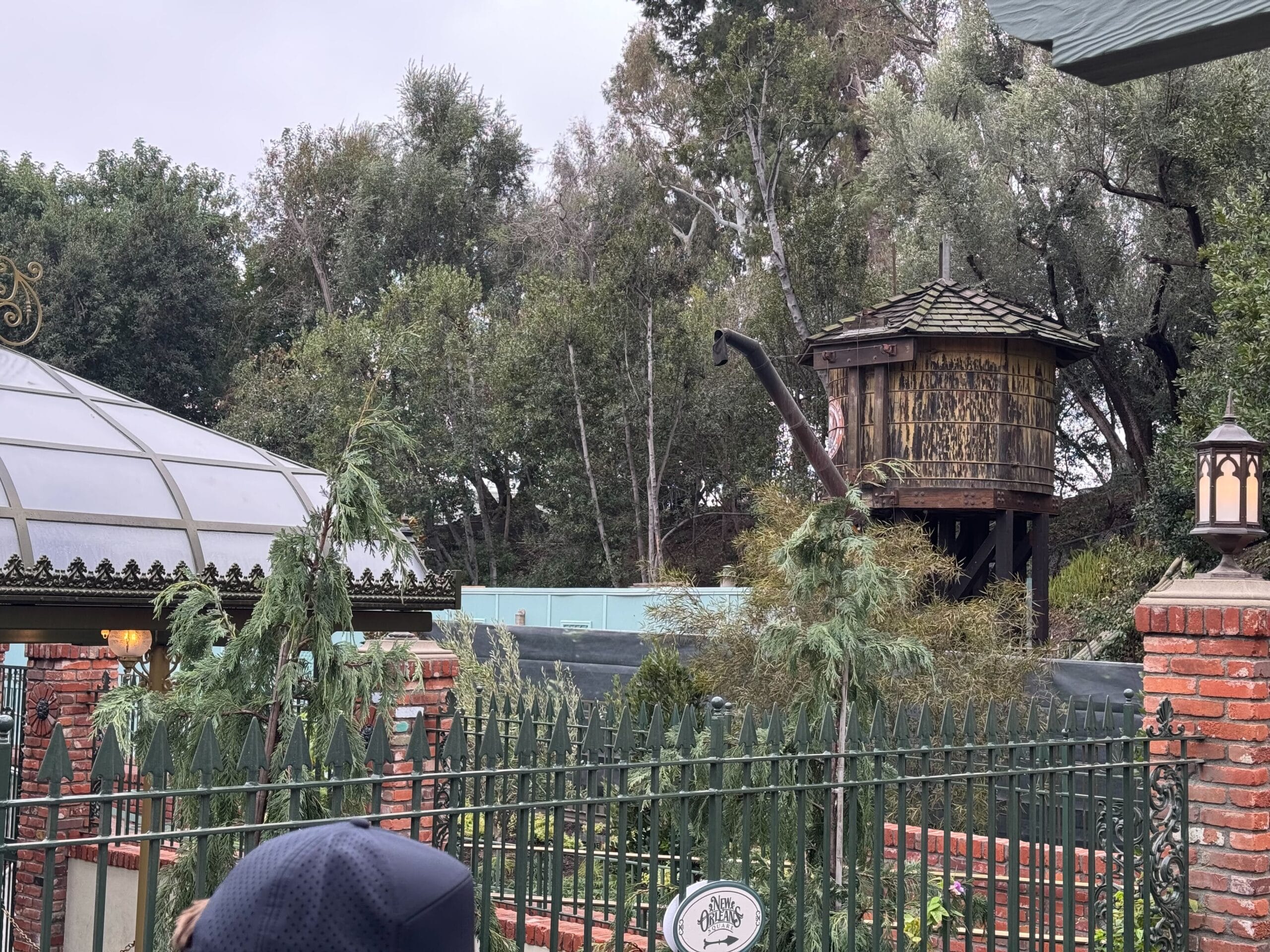 An outdoor scene featuring a water tower and greenhouse roof alongside brick structures evokes a quaint charm, reminiscent of the architectural style found in New Orleans Square. The setting is enveloped by trees and bordered by a dark metal fence, adding to its secluded allure.