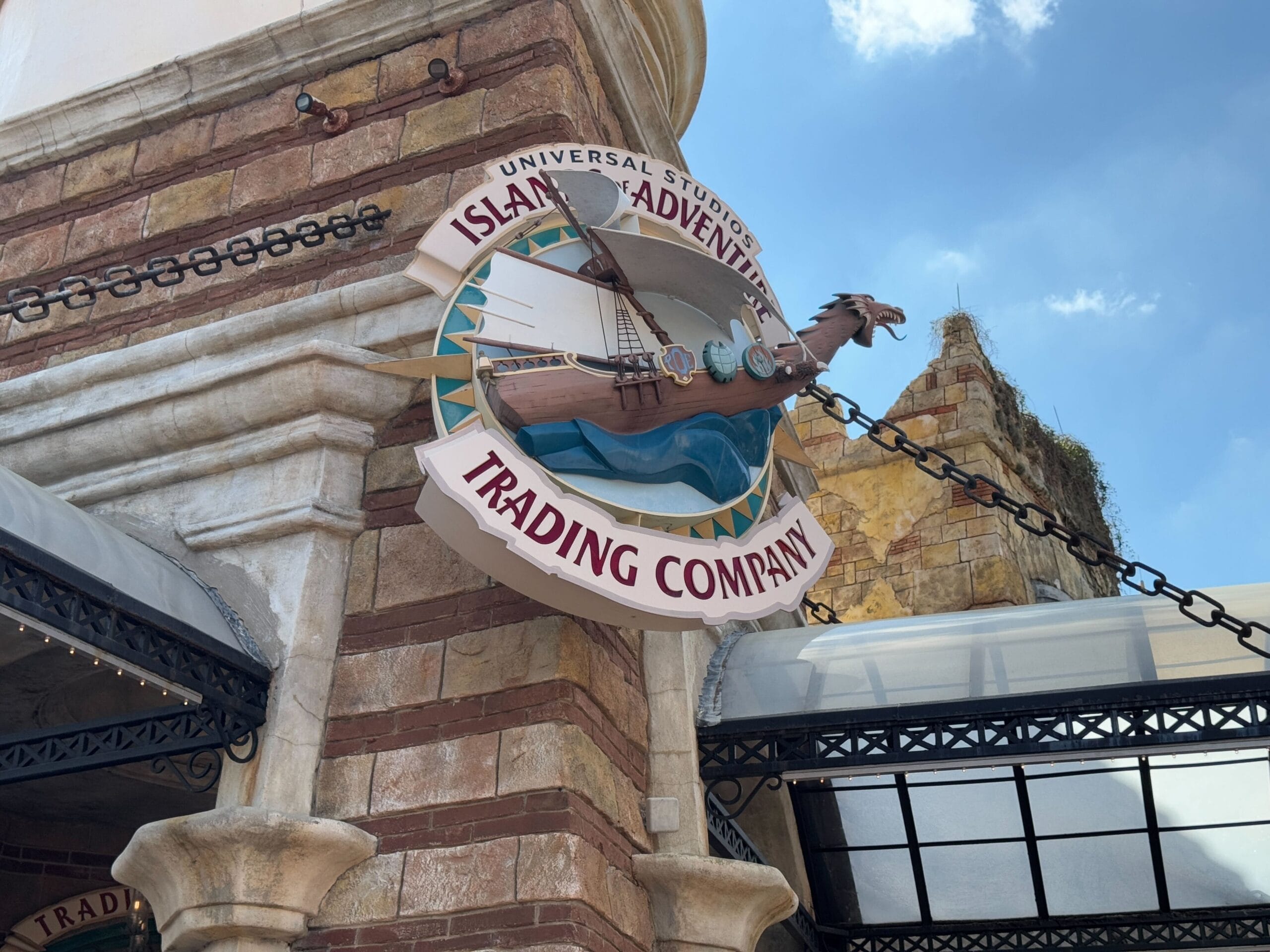 Sign for "Islands of Adventure Trading Company" with a ship illustration, mounted on a brick and stone wall, offering a hint of the epic universe beyond through the partial view of building structures in the background.