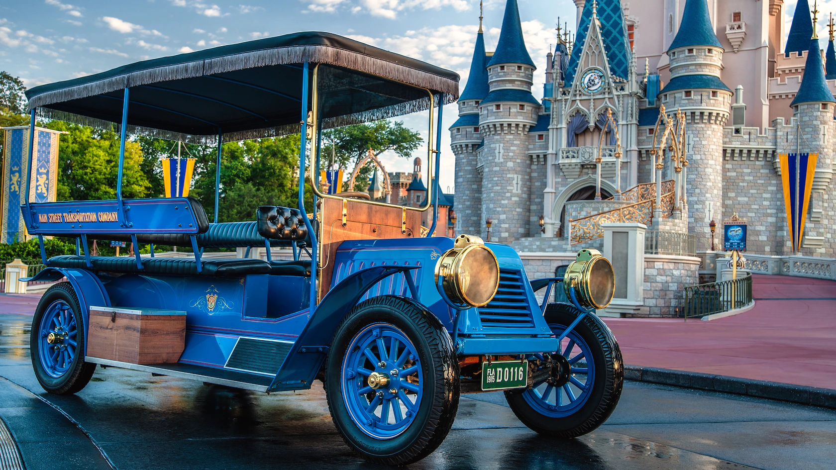A vintage blue car from the Main Street Vehicles collection is parked on a wet street in front of a fairytale castle, showcasing its blue turrets and decorative flags.