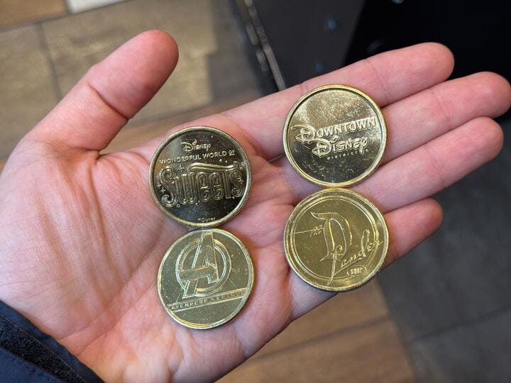 A hand proudly displays four golden tokens, each a unique collectible medallion showcasing intricate designs, including the iconic "Downtown Disney" and "Avengers Reserve" logos.