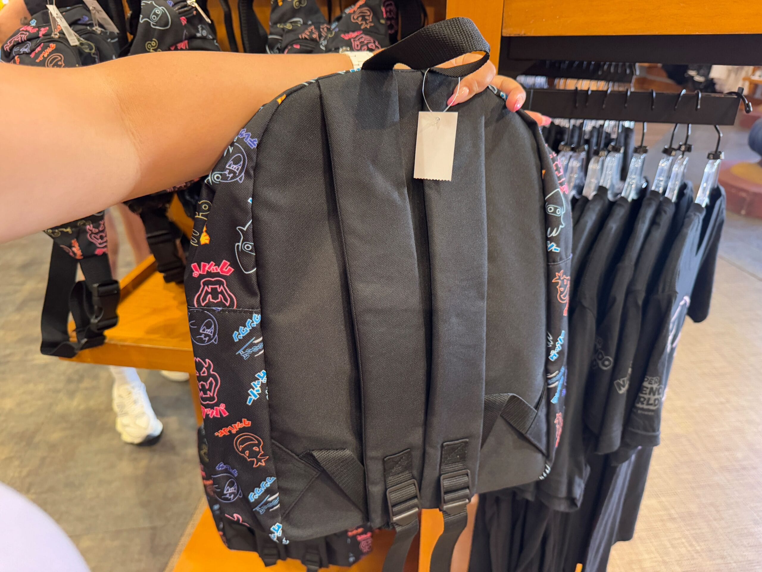 A person holds a black backpack adorned with colorful outlines of cartoon faces, an eye-catching piece from the Epic Universe Merchandise collection. It hangs beside black t-shirts on display, capturing the vibrant energy of Florida's Universal Studios.