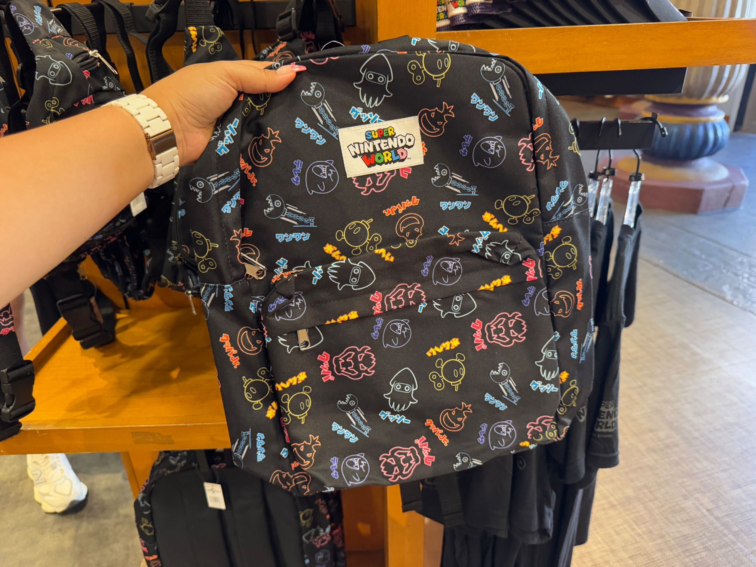 A hand holds a black backpack with colorful cartoon characters and "Nintendo World" text, showcased on a wooden shelf alongside Epic Universe Merchandise from Universal Studios Florida.