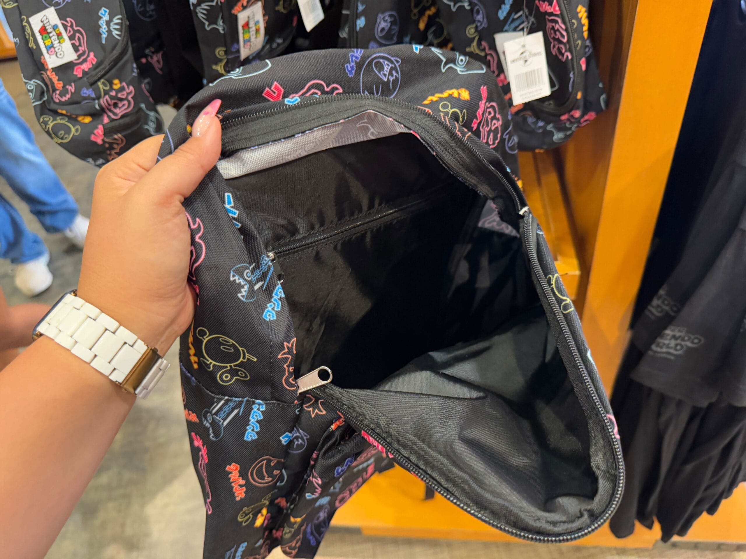 A person holds an open black backpack adorned with colorful cartoon doodle designs, revealing its interior—a perfect piece of Universal Studios Florida's Epic Universe merchandise. Similar vibrant backpacks are artfully displayed in the background.