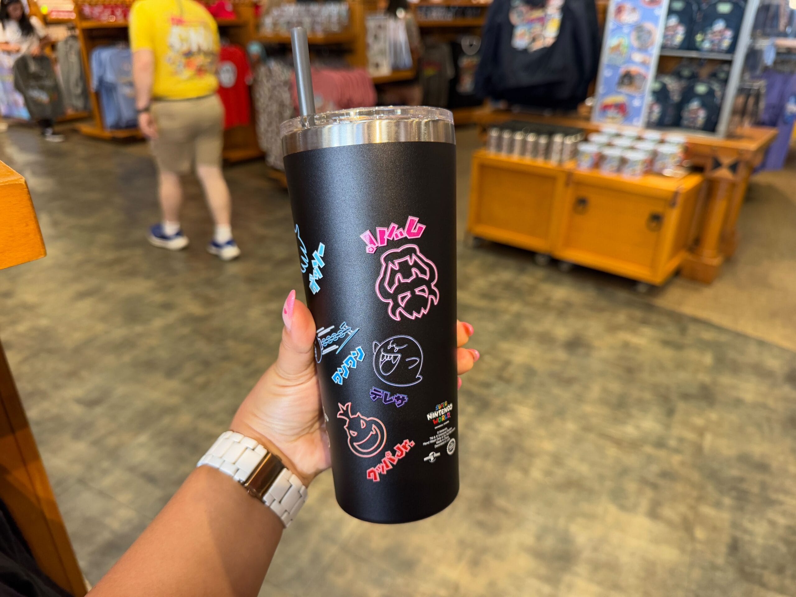 A person holds a black tumbler adorned with colorful graphics representing Epic Universe Merchandise, featuring a straw, inside a store with wooden shelves and a blurred background.