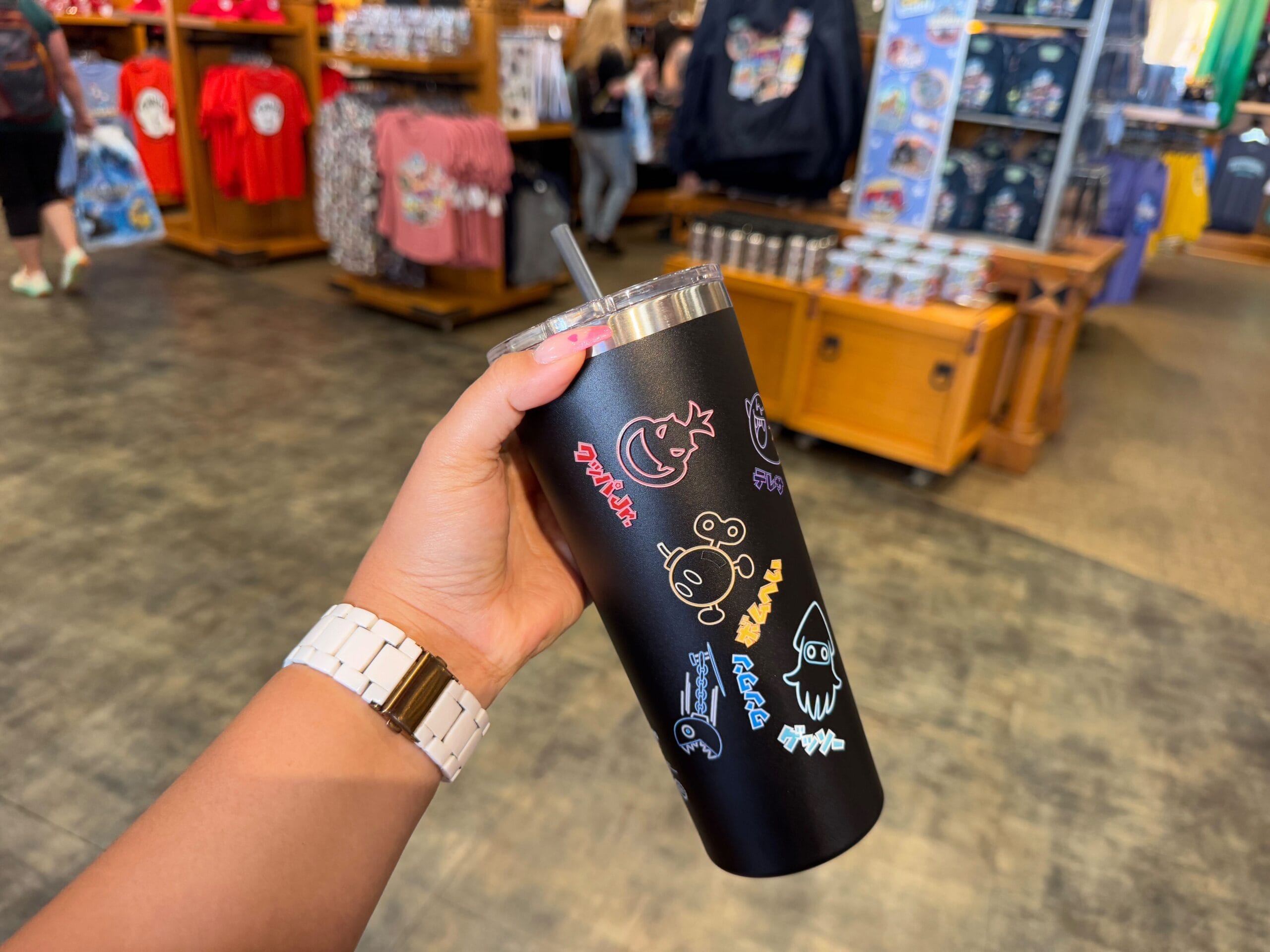 A hand holds a black tumbler adorned with colorful cartoon designs, set against the backdrop of a store full of Epic Universe Merchandise at Universal Studios Florida. Shelves brim with clothing and trinkets, creating a vibrant shopping paradise for all fans.