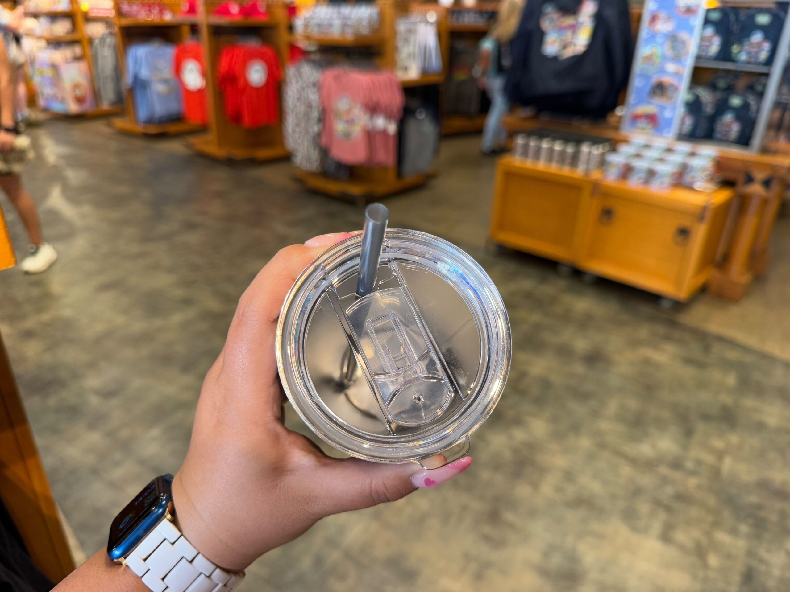 A hand holds a clear lidded cup with a straw, set against the vibrant backdrop of Epic Universe Merchandise in a bustling retail store, where new merchandise and shirts are proudly on display.