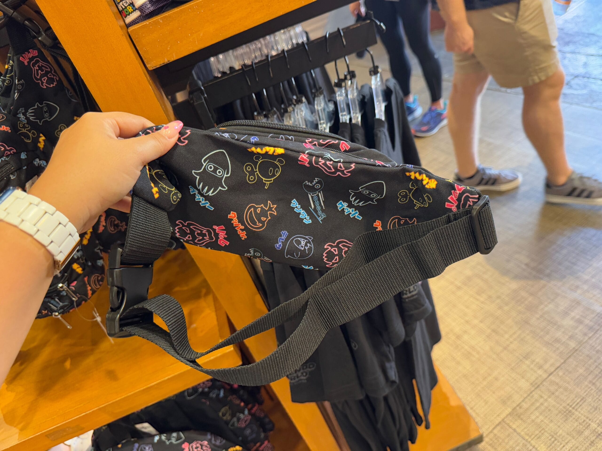 A person holds a black belt bag with colorful cartoon designs in a store, showcasing the NEW Epic Universe Merchandise from Universal Studios Florida.