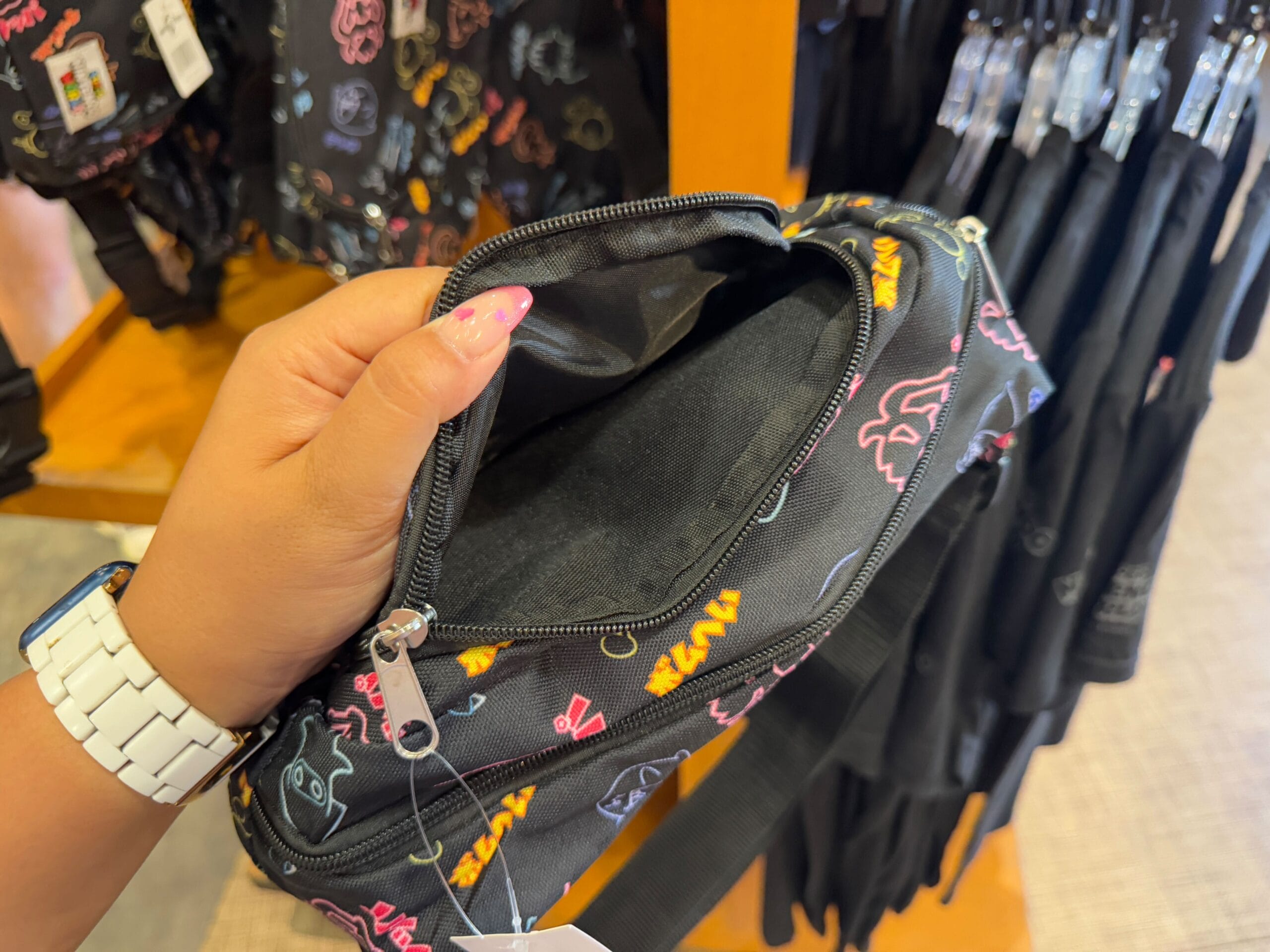 A person holds an open black bag adorned with colorful line art designs, reminiscent of Epic Universe Merchandise. The bag's empty interior is visible as the individual flaunts a white watch and pink nail polish, perhaps ready for a day at Universal Studios Florida.