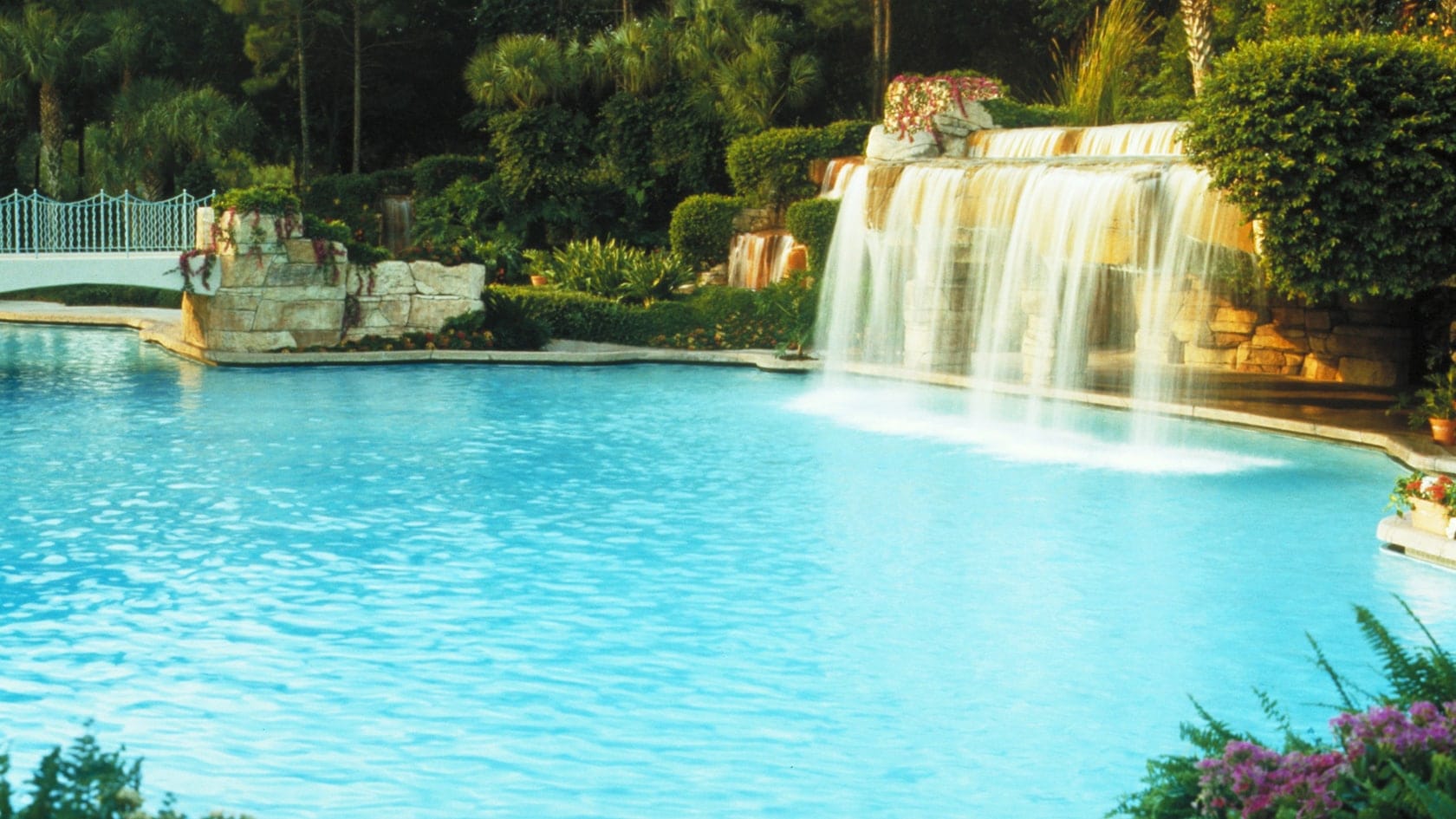 A large swimming pool with a waterfall at Splash Terrace, surrounded by lush greenery and a white decorative bridge.